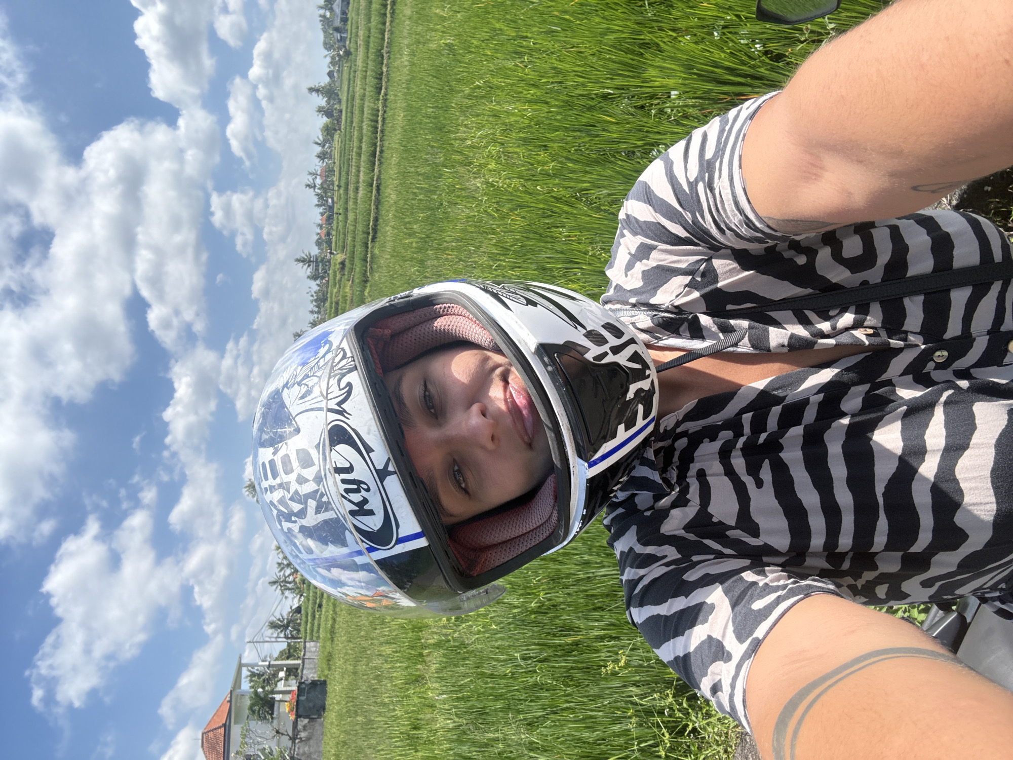 A first-person shot depicts a person wearing a motorcycle helmet and a zebra-striped shirt, seemingly on a motorcycle, with a verdant rice field forming the backdrop. The sky is visible, exhibiting a mix of clear blue and fluffy white clouds. A glimpse of a building structure is evident in the distance. The location is Pejeng, Indonesia, identifiable by the distinctive rice terraces.