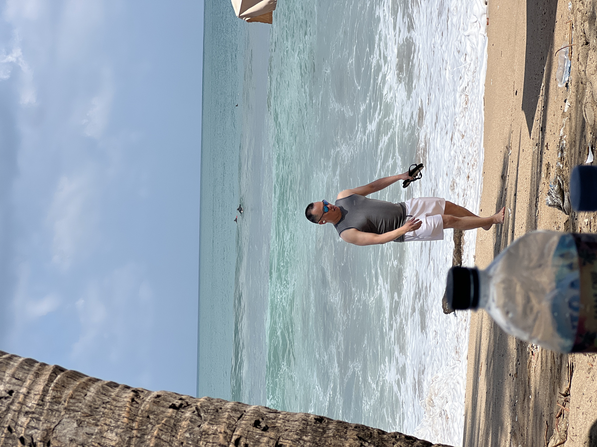 In Kuta, Indonesia, a man walks along a sandy beach with the ocean to his right. He is wearing a grey tank top and white shorts, and he carries a pair of black flip-flops in his right hand. The ocean is a turquoise color with white waves crashing on the shore. There are at least three dolphins visible in the distance. In the foreground, a clear plastic bottle with a black lid sits on the sand. To the left, the rough bark of a palm tree trunk partially obstructs the view. The sky is a pale blue with scattered clouds.