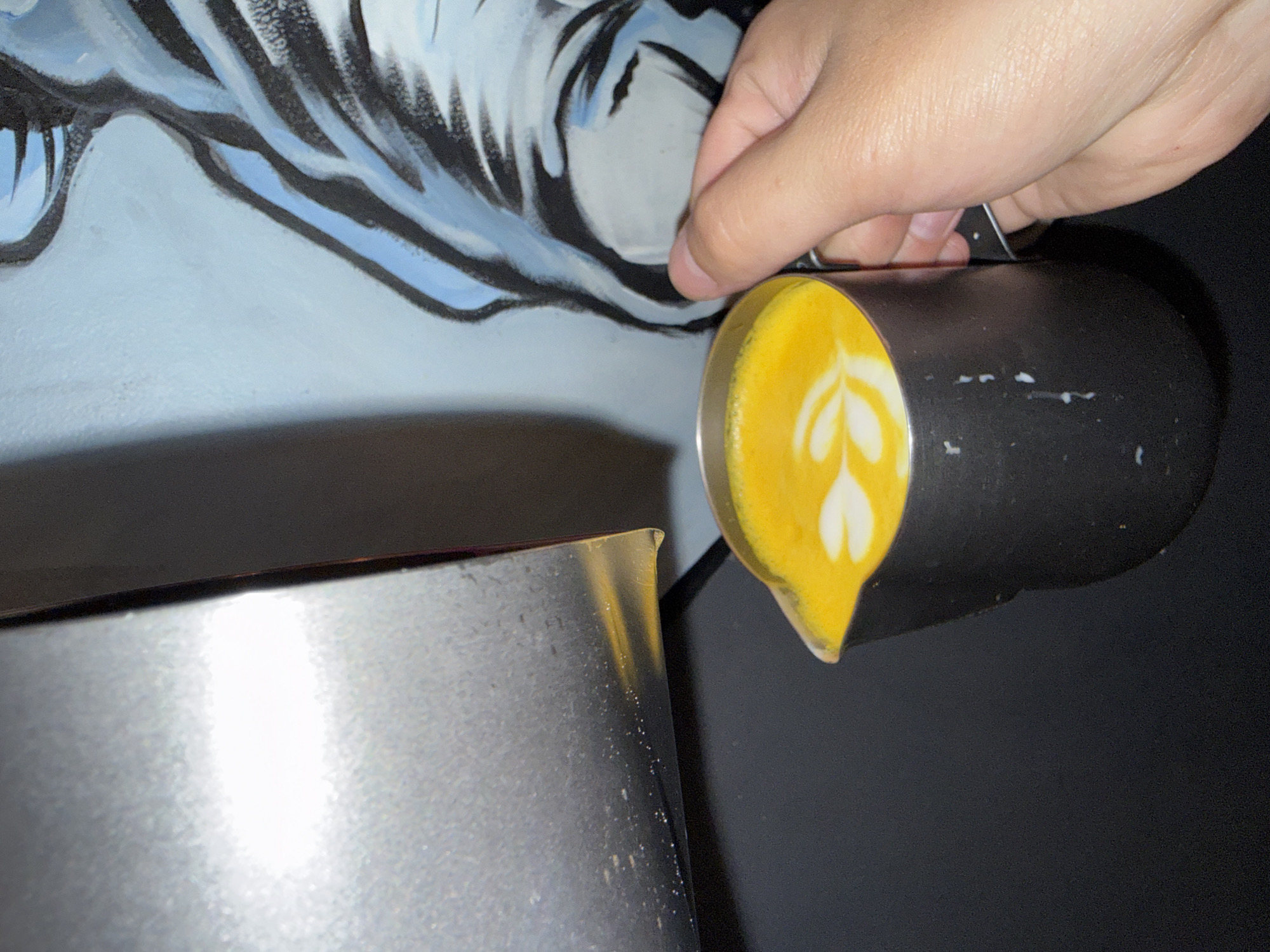A close-up, angled shot captures the moment a hand pours a yellow-colored beverage with latte art from a black metal pitcher into a larger, silver metal container. The hand, with light skin tone, holds the handle of the black pitcher, tilting it downwards to release the liquid. The surface of the yellow liquid in the black pitcher displays a distinct white latte art pattern, resembling a stylized tulip or a series of hearts. Small white splatters are visible on the exterior of the black pitcher, suggesting prior use or spillage.
Partially visible in the background is a wall adorned with a painted mural. The mural features abstract or stylized shapes in white and light blue, outlined with bold black strokes, possibly depicting a face or animal-like features, or simply decorative brushwork. The area behind the silver container is dark, contrasting with the lighter, artistic wall. The silver container itself is reflective, showing a bright highlight. The scene suggests an indoor setting, likely a cafe or a preparation area, in Khimki, Russia, focused on the art of beverage making. No visible text is present in the image.