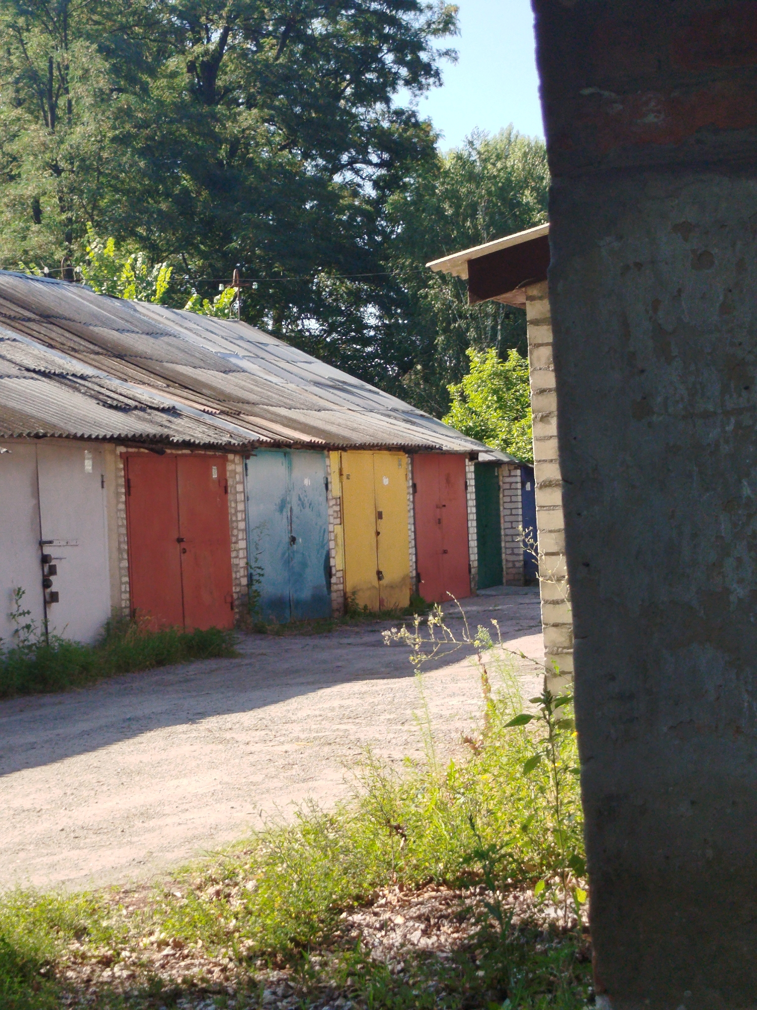 In Kremenchuk, Ukraine, a row of garages with corrugated metal roofs lines a dirt road. The garages have various colored doors, including white, red, blue, and yellow. Vegetation, including grass and small trees, grows around the garages and on the roadside. A large, unpainted concrete structure partially obstructs the view on the right side of the image, suggesting it's a neighboring building or a part of another garage complex. Trees and foliage form a backdrop against a clear blue sky, indicating an outdoor, possibly suburban or industrial, setting. The overall scene depicts a utilitarian area, likely a garage cooperative or storage facility, under bright daylight.