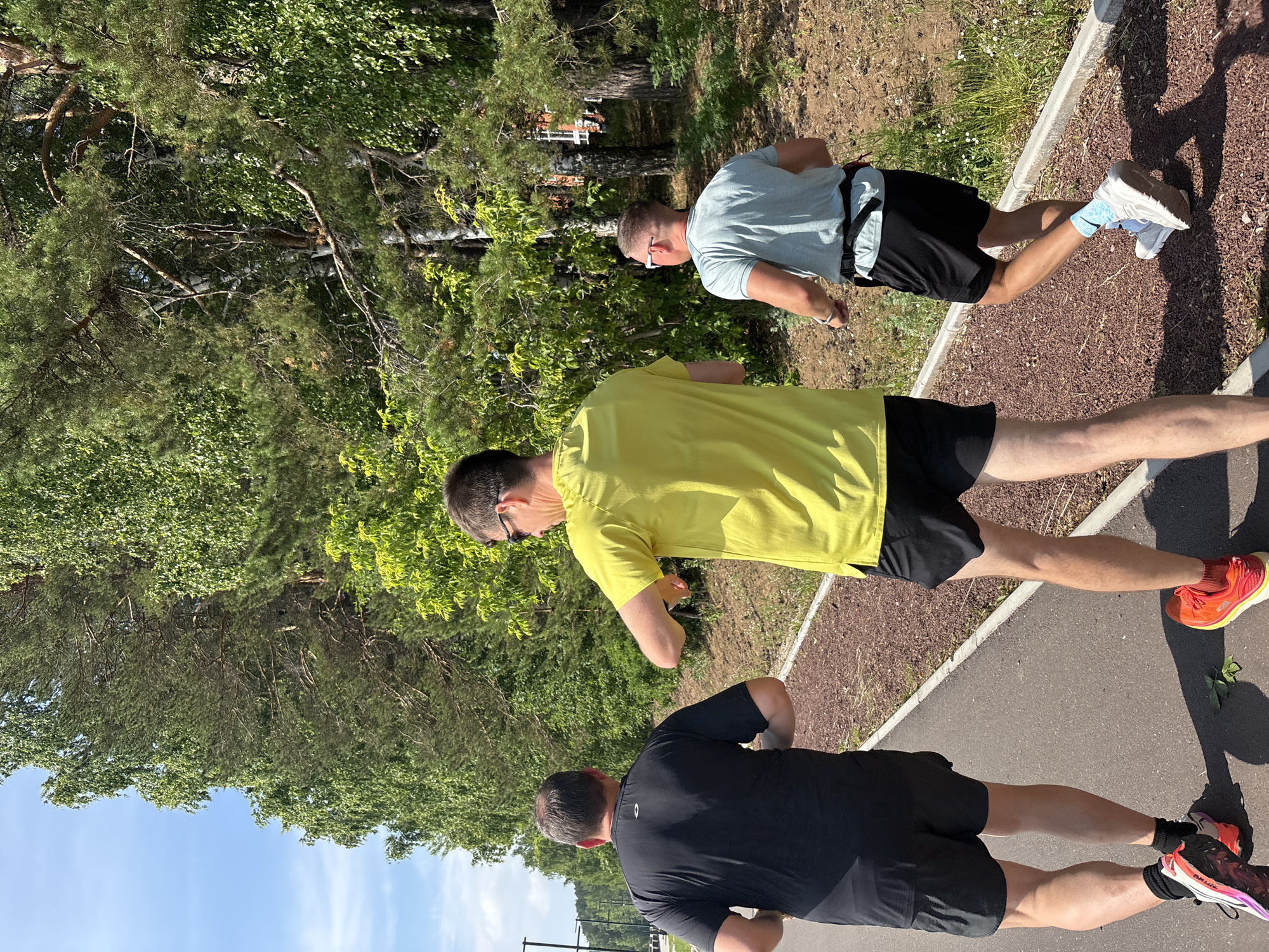 The image depicts three men running on a paved path adjacent to a wooded area. The path appears to be a dedicated running or cycling trail, with a dark grey asphalt surface and a reddish-brown gravel border on the right side. The wooded area consists of tall trees, predominantly coniferous with some deciduous trees interspersed, suggesting a park or a natural reserve setting within or near a city. The sky is blue with some scattered clouds, indicating fair weather.
From left to right, the first man is wearing a black t-shirt and black shorts, and he is viewed from behind. He has athletic build and is wearing colorful running shoes. The second man, in the center, is wearing a bright yellow t-shirt and black shorts. He is also viewed from behind and appears to be mid-stride, with his left arm raised and bent. He is wearing orange running shoes. The third man, on the right, is wearing a light blue t-shirt and black shorts. He is viewed from the back and side, and he is wearing white and grey running shoes with light blue socks. He has sunglasses on. He is also running.
The overall environment is green and well-maintained, suggesting an outdoor recreational space. The presence of running trails and the attire of the individuals strongly indicate a physical activity context, likely a morning or afternoon jog. The lighting suggests it's daytime, with sunlight casting shadows on the path.
The image can be located in the city of Chaikovsky, Russia. The specific location is likely a park or a recreational area within the city, such as the Central Park or along the Kama River embankment, which are known for their green spaces and outdoor activity facilities. The type of trees and the general landscape are consistent with the natural environment of the Perm Krai region, where Chaikovsky is situated.