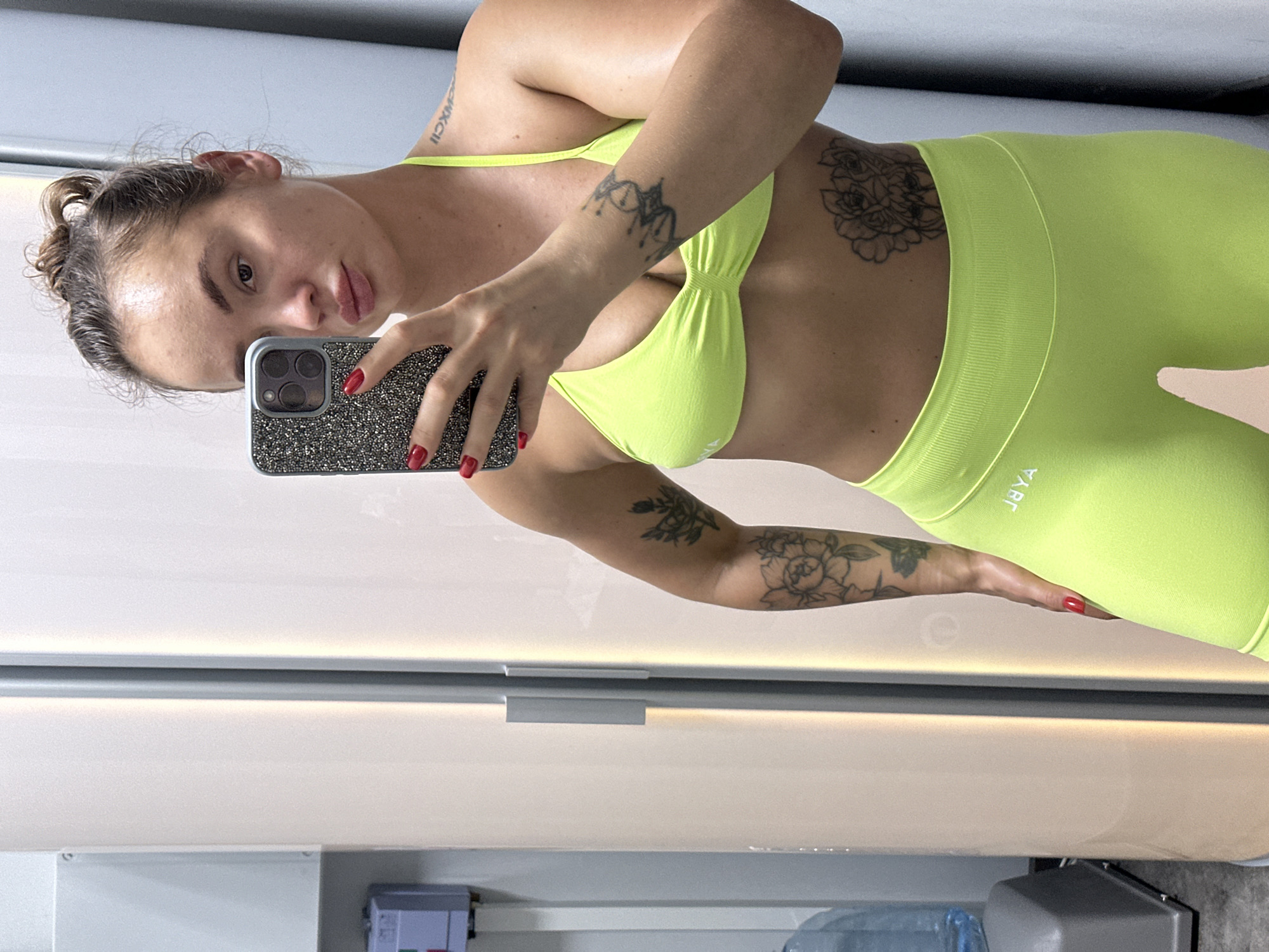 A woman with her hair pulled up in a bun takes a selfie in a gym locker room. She is wearing a neon yellow sports bra and shorts. She has tattoos on her arms and torso. A water cooler and locker are visible in the background.