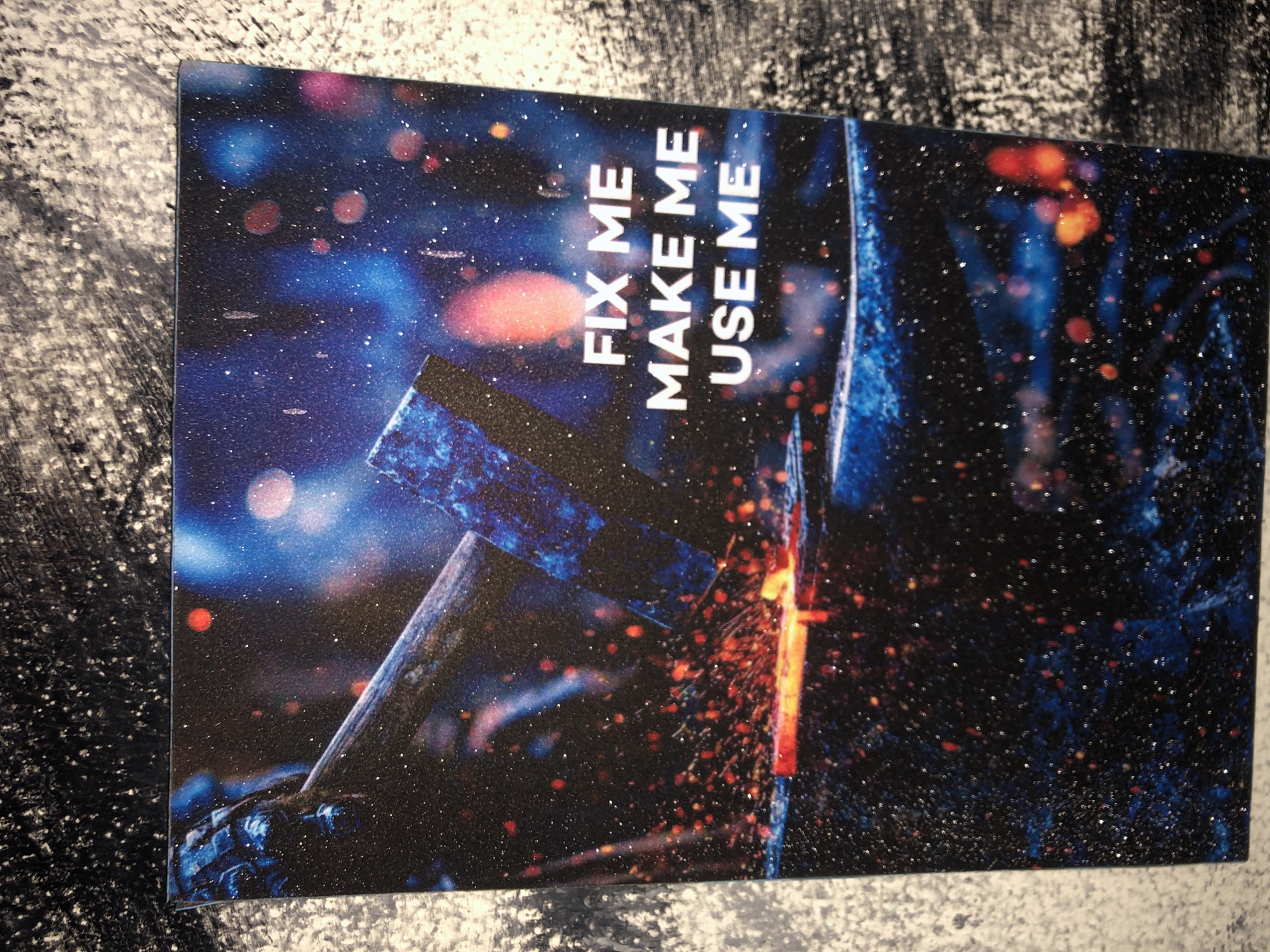 A close-up, overhead shot of a poster with a glittery, textured surface is shown. The poster depicts a blacksmith's anvil with a glowing red-hot piece of metal on it. A large hammer, made of dark, reflective material, is poised to strike the metal, sending sparks flying. The background is a dark, starry, and cosmic-looking scene with out-of-focus blue and red lights. Text on the poster reads "FIX ME MAKE ME USE ME" in white block letters. The poster is placed on a textured, black and white surface, possibly a wall or backdrop, which has a rough, painted appearance with visible brushstrokes. The overall effect is dramatic and emphasizes the act of creation and transformation. The location in Kabong, Nigeria is not specified or visible in the image.