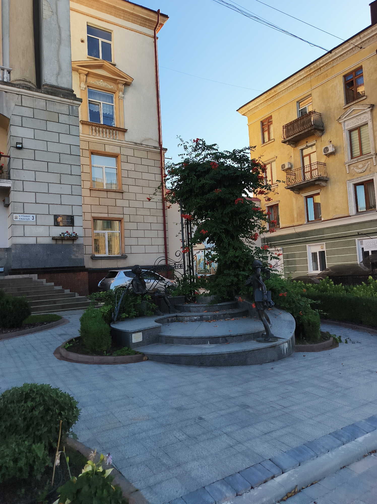 The image shows a street scene in Ternopil, Ukraine. In the foreground, there is a paved area with two small bronze statues of children. One statue depicts a child sitting on a bench, and the other shows a child holding a book. These sculptures are part of a public art installation. Behind the statues, there is a circular arrangement of steps and a lush green tree covered in red flowers. The background features two buildings. The building on the left is lighter in color with multiple windows and a stone facade. A street sign on this building indicates "вул. М. Грушевського, 3" which translates to "Hrushevsky Street, 3". The building on the right is yellow with a greenish lower level and features balconies with ornate railings. The scene is illuminated by natural light, suggesting it is daytime. The environment appears to be a well-maintained urban public space.