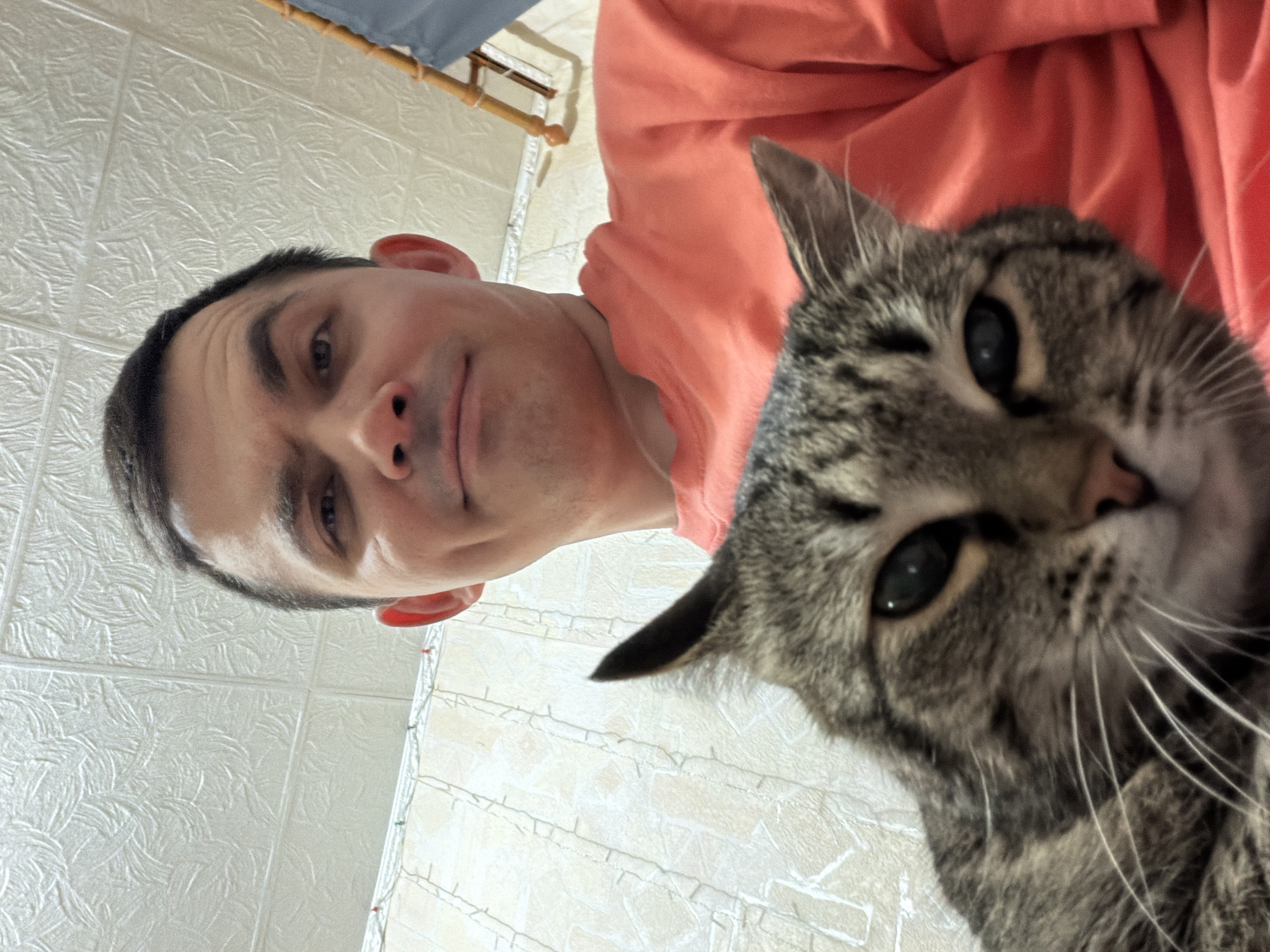 A selfie-style image shows a man and a cat indoors. The man, with short dark hair and light skin, is wearing a peach-colored shirt. The cat, a tabby, is positioned close to the camera, its face filling much of the frame. The background is a white wall with a textured pattern. The image's angle is tilted. The location appears to be indoors. There is no indication of Izhevsk, Russia.