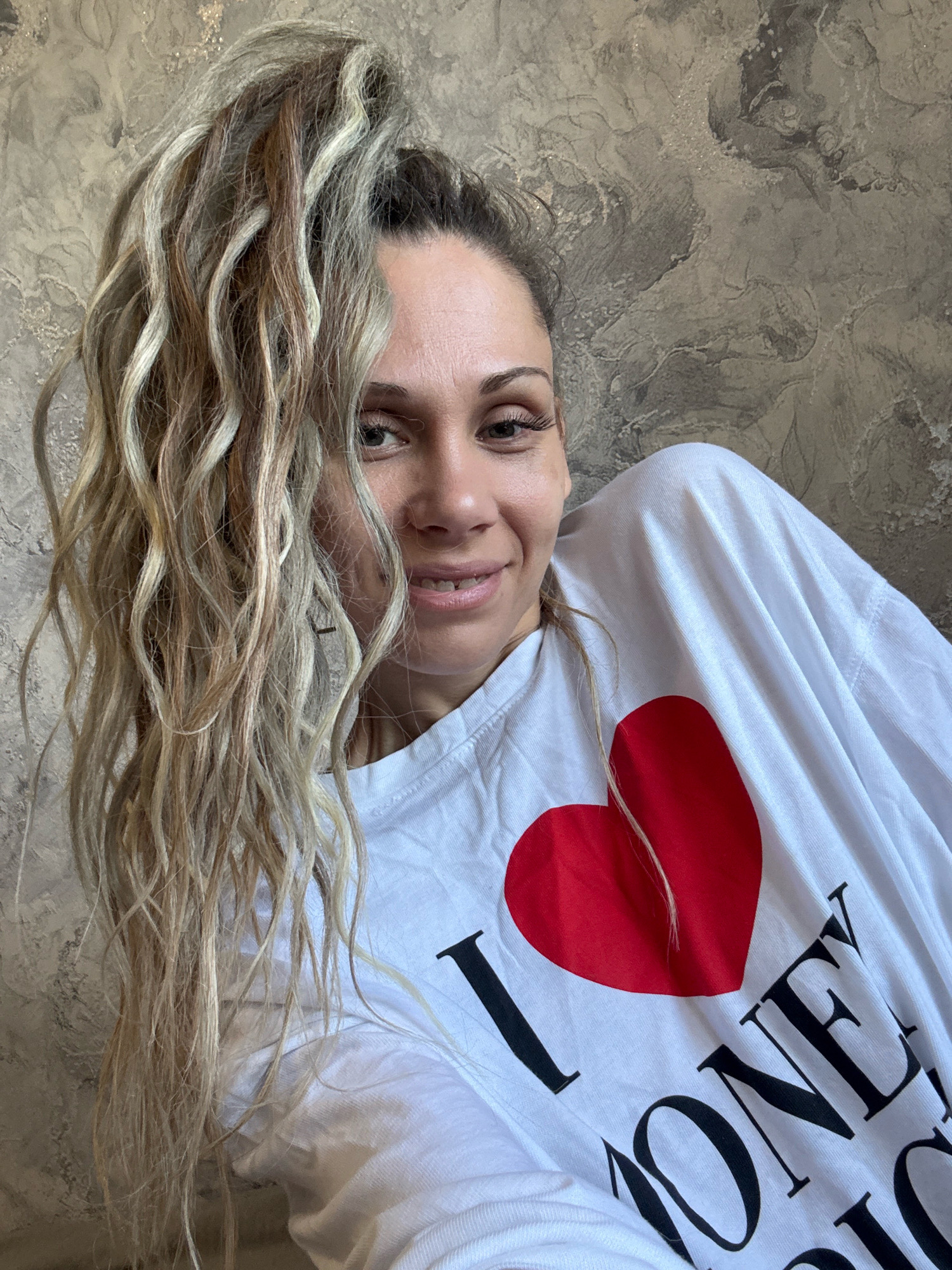 A woman with blonde, wavy hair pulled back in a high ponytail is shown from the shoulders up, leaning to her right with her head tilted back. She is wearing a white t-shirt with a large red heart and text that reads "I LOVE MONEY" in black. Her eyes are open and she is smiling. The background is a textured gray wall. The image was taken in Rusanovo, Russia.