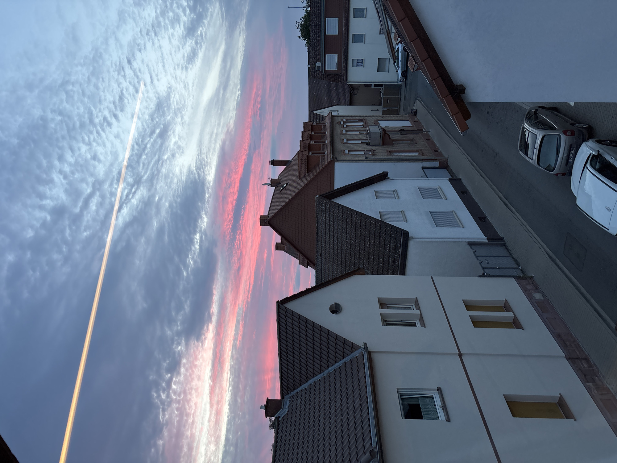 The image depicts a residential street scene in Viernheim, Germany, at dusk. The sky is a dramatic canvas of pink and gray clouds, illuminated by the setting sun, with a bright contrail streaking across the upper portion of the frame. Row houses with tiled roofs line the street, exhibiting various architectural styles. The facades of the houses are predominantly white, with some featuring red-tiled roofs and dormer windows. Two cars are parked on the street: a silver hatchback and a white sedan. The street is paved with cobblestones, and a sidewalk is visible. A metal fence borders one of the properties. The overall impression is one of a quiet, European neighborhood as daylight fades.