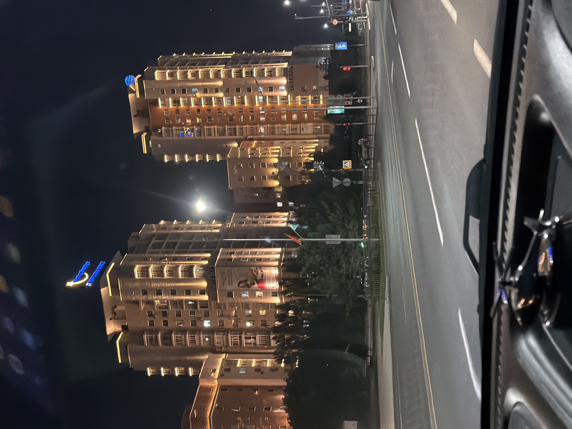 The image captures a nighttime view of modern, illuminated residential buildings in Astana, Kazakhstan. The buildings are tall and multi-story, with warm yellow lights highlighting their architectural features. One building prominently displays a blue neon sign on its rooftop, forming a crescent moon shape with Arabic script below. Another adjacent building has a large blue sphere on its roof. The foreground shows a street with traffic lights and trees. In the immediate foreground, the dashboard of a vehicle is visible, with a small reflective object, possibly a dashboard ornament, in the center. The streetlights and building lights create a bright, urban atmosphere against the dark night sky.