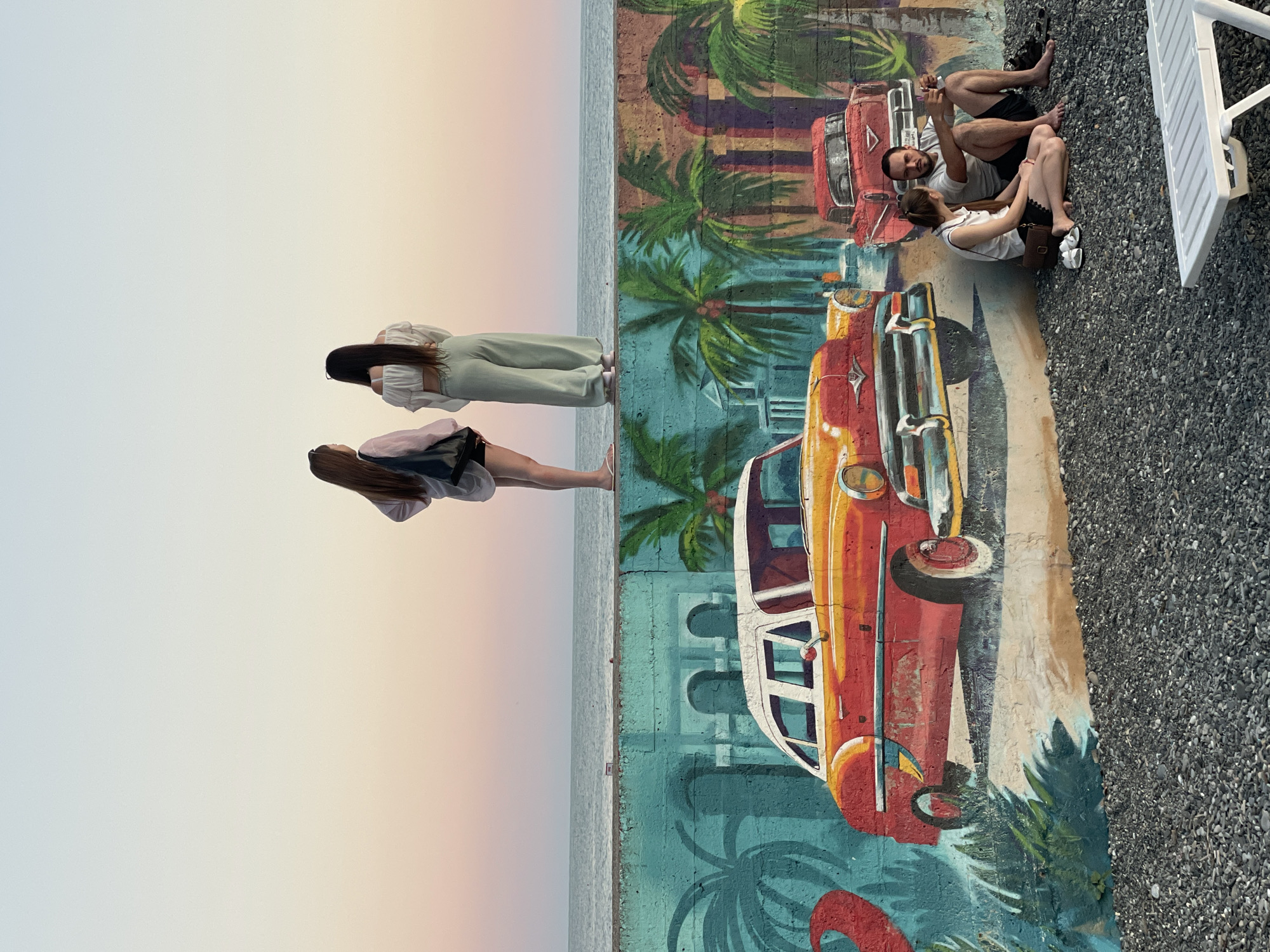 Two women stand on a sea wall, silhouetted against a pale pink and blue sunset sky. Below them, a mural of palm trees and vintage cars adorns the wall. A couple sits on the pebbled ground in front of the mural, the man holding a phone as if taking a selfie. A white lounge chair sits to the right, partially in the frame. The scene appears to be a seaside promenade in Sochi, Russia.
