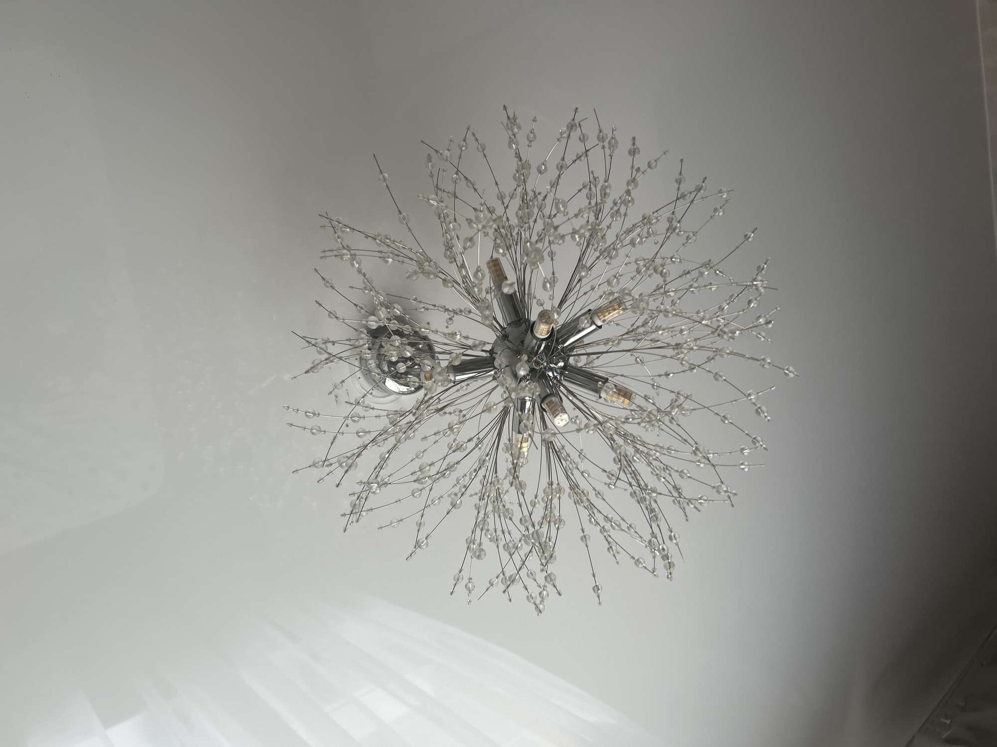 A modern chandelier with a chrome base and thin, wire-like extensions adorned with clear, crystalline beads hangs from a white ceiling. Several small, orange-tinted light bulbs are visible among the beads. Sunlight casts a pattern of diffused light and shadows on the ceiling, particularly on the left side of the image, suggesting the presence of a window with sheer curtains. The overall impression is one of a well-lit interior space, likely a room or hallway. The image does not provide enough information to determine its specific location in Saint Petersburg, Russia, or the presence of any individuals.