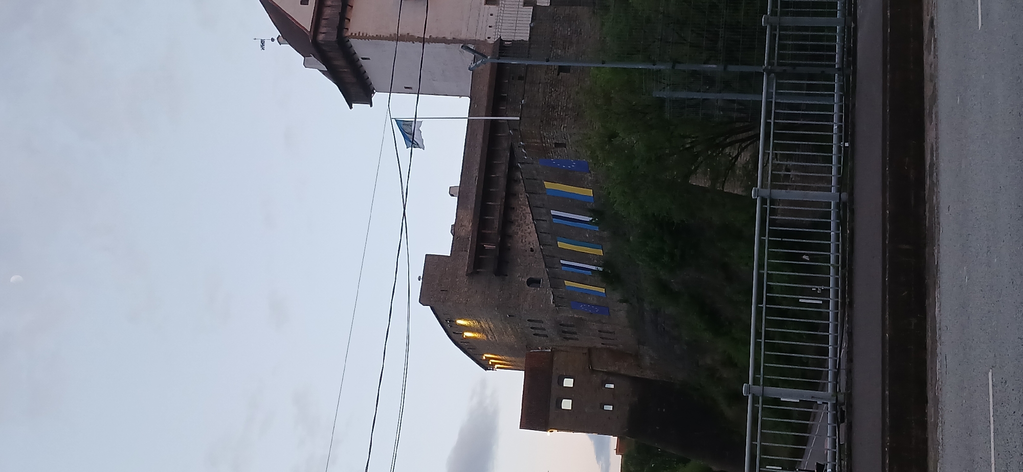 The image captures a view of Ivangorod Fortress in Russia, situated alongside a street and bridge railing. The fortress is constructed of dark stone, with a tall, imposing structure punctuated by windows. Flags are displayed along the fortress wall, including what appears to be Ukrainian flags and EU flags. Overhead, power lines stretch across the sky, which is light, suggesting twilight or early evening. A building stands adjacent to the fortress, and greenery is visible around the base of the structure. The road next to the railing and the overcast sky indicate a relatively cool or temperate weather condition.