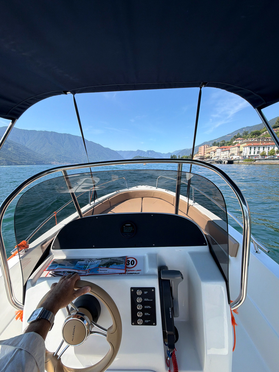 A first-person perspective shot from a boat on a body of water, likely Lake Como, near the city of Tremezzina, Italy. The scene is dominated by a blue sky and distant, verdant mountains. In the foreground, a white boat's interior is visible, featuring a beige-upholstered seat, a steering wheel with the "ALLEGRA" logo, a throttle control, and a dashboard with various buttons and a digital display. A hand wearing a wristwatch is seen gripping the steering wheel. A speed limit sign indicating "30" and "KM/H SPEED LIMIT" is attached to the dashboard. The water is a deep blue, reflecting the sky and mountains. Buildings with terracotta roofs are visible along the distant shoreline.