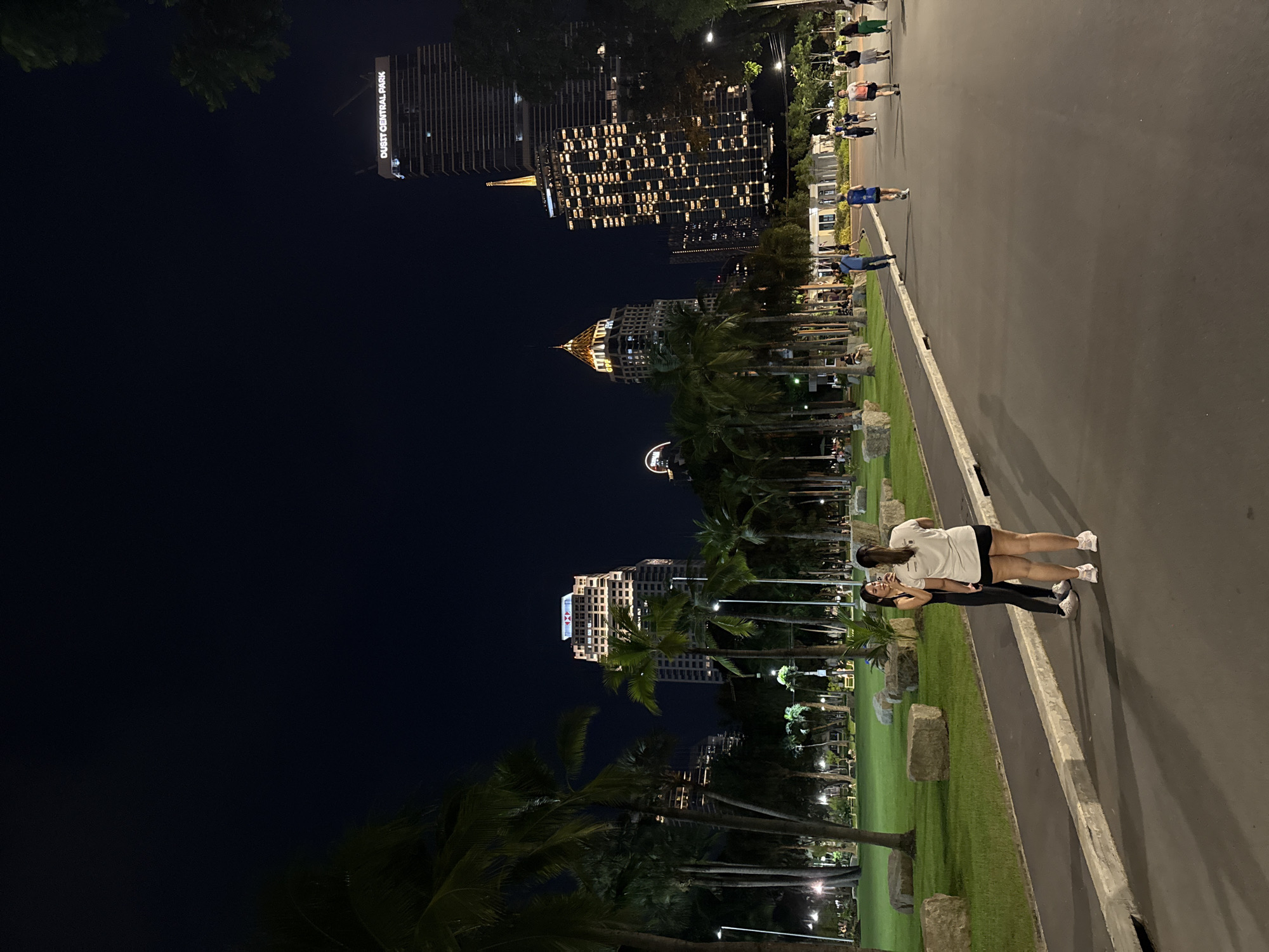 At night in Bangkok, Thailand, two women stand on a walkway next to a grassy area with palm trees. The women appear to be young adults. One wears a white shirt and black shorts; the other wears a black top and black leggings. The walkway is paved, and a concrete barrier separates it from the grass. In the background, several illuminated buildings dominate the skyline. One building is labeled "Dusit Central Park." People walk along a sidewalk in the background. The sky is dark.
