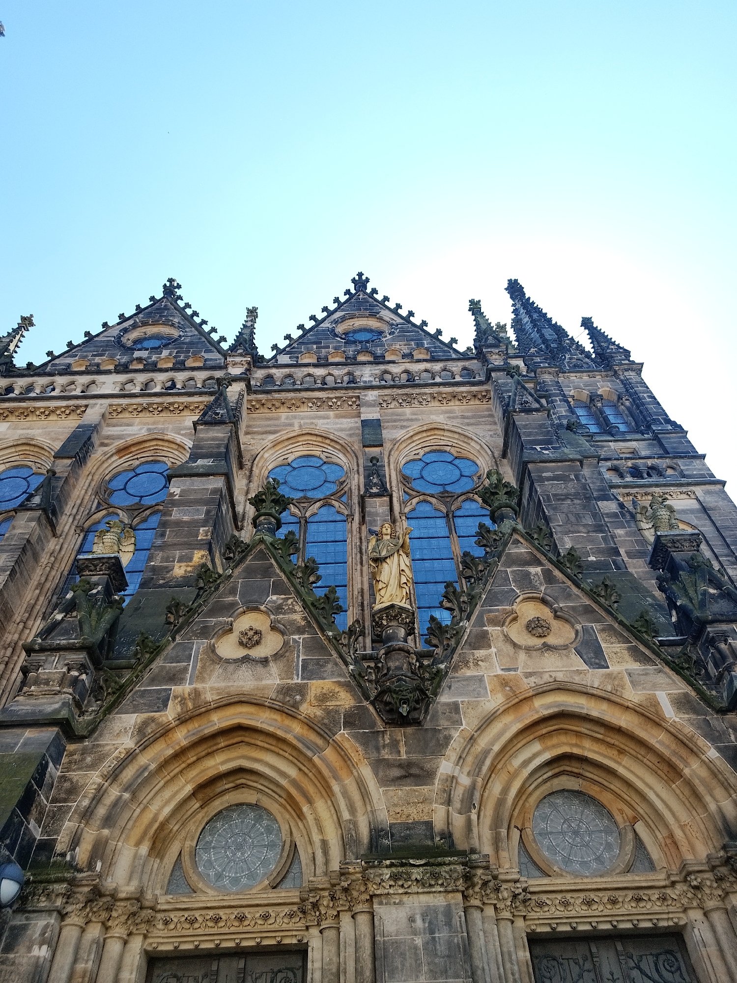 The image shows a low-angle shot of a large, ornate stone building in the city of Unknown, Unknown. The building appears to be a church or cathedral, with arched windows, intricate stone carvings, and multiple spires. A statue stands above the entrance. The sky is visible at the top of the image and is a clear blue.