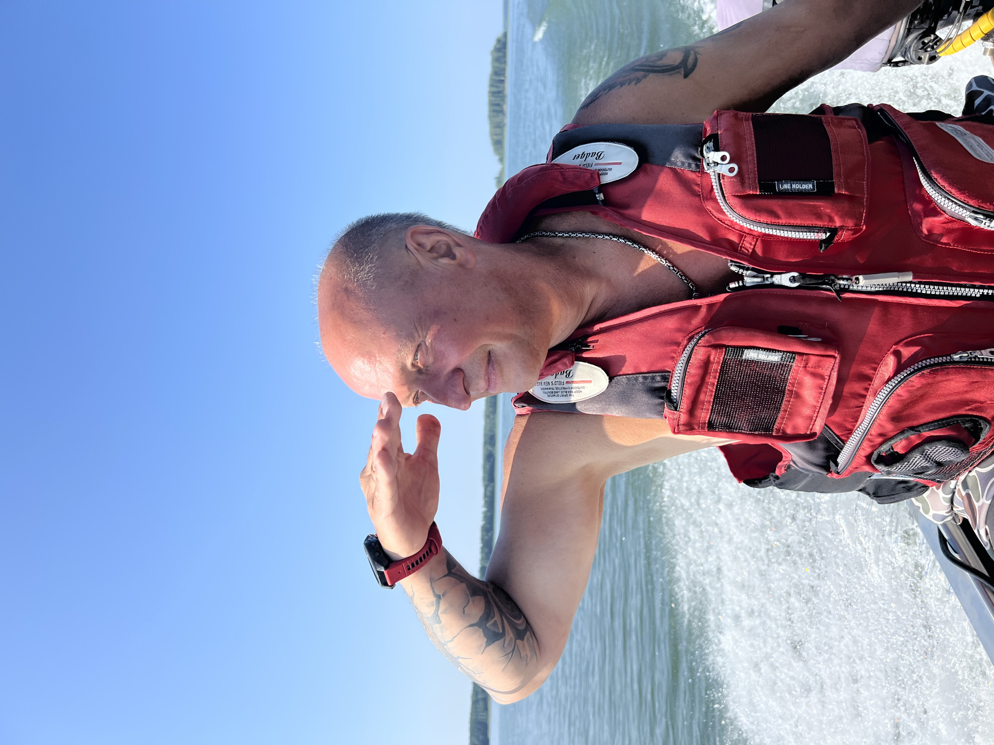 A bald, fair-skinned man in his 50s with a tattoo on his left arm and a silver chain around his neck is operating a motorboat on a body of water. He is wearing a red life vest with multiple pockets and zippers. He is looking out at the horizon with his right hand shielding his eyes. The sky is clear and blue, and there is a wake behind the boat. In the distance, there are trees on an island. The location is the city of Beregovoe, Russia.