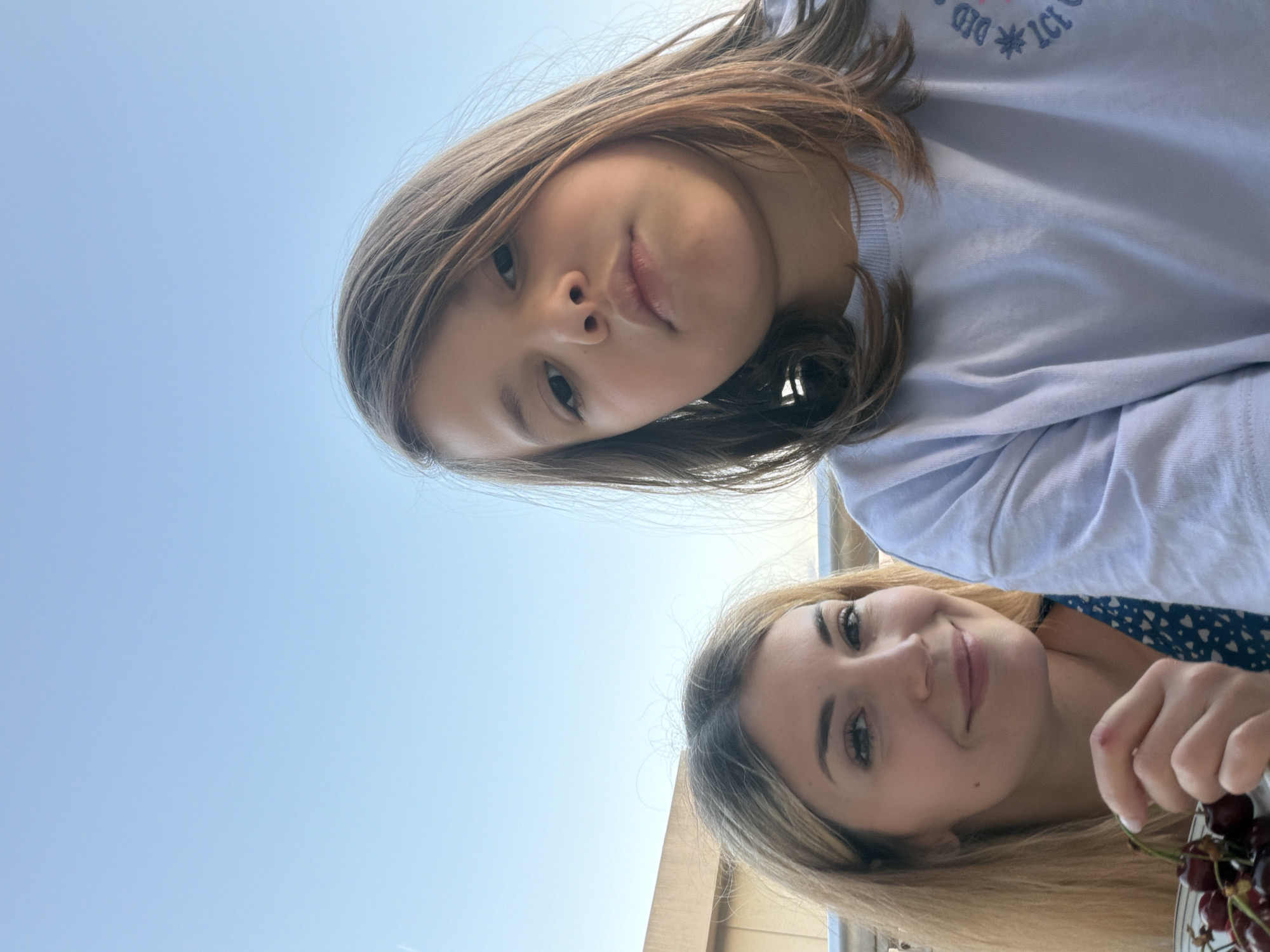 A selfie-style photograph shows a young girl and an adult woman. The girl has brown hair, brown eyes, and is wearing a light blue t-shirt with lettering. The woman has blond hair, light skin, and is holding a plate of dark red cherries. The background consists of a light blue sky and a light-colored building. The photograph appears to be taken from a high angle, looking down towards the subjects. The location cannot be determined as being in Budva, Montenegro with the current information.