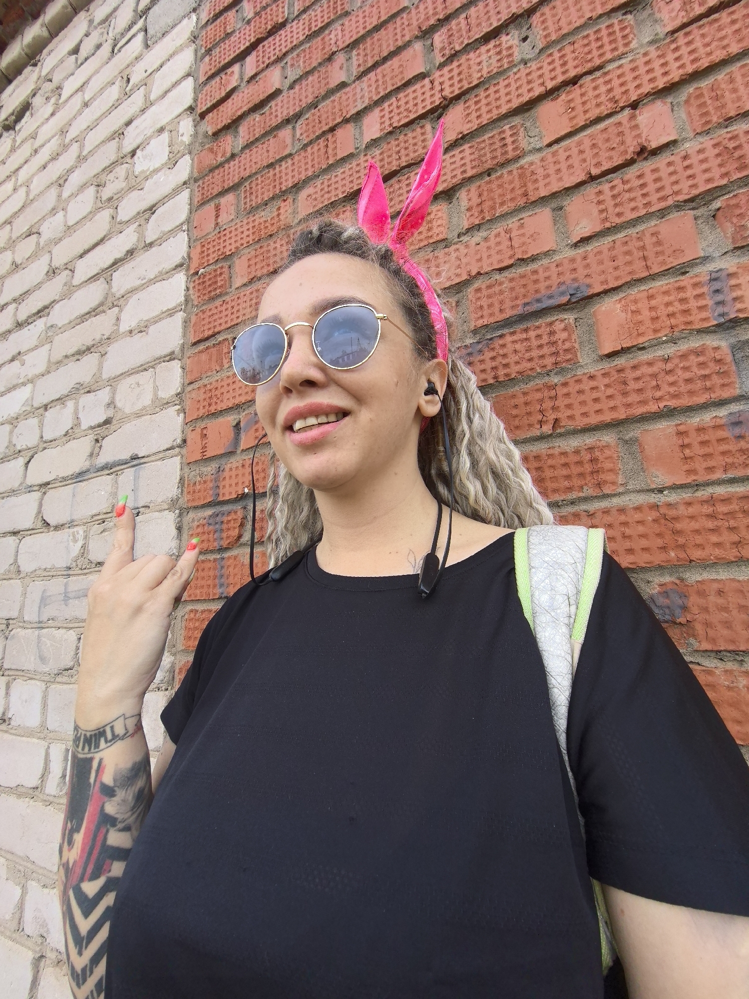 A woman stands outdoors against a two-tone brick wall, one side whitewashed and the other red brick, possibly in Noviy, Russia. She wears round, gold-rimmed sunglasses, a pink bunny ear headband, and a black t-shirt. A grey backpack with green trim is slung over her shoulder. She has wireless earbuds. Her left arm displays a full sleeve tattoo, and she makes a "rock on" hand gesture. Her fingernails are painted with a green-orange gradient.