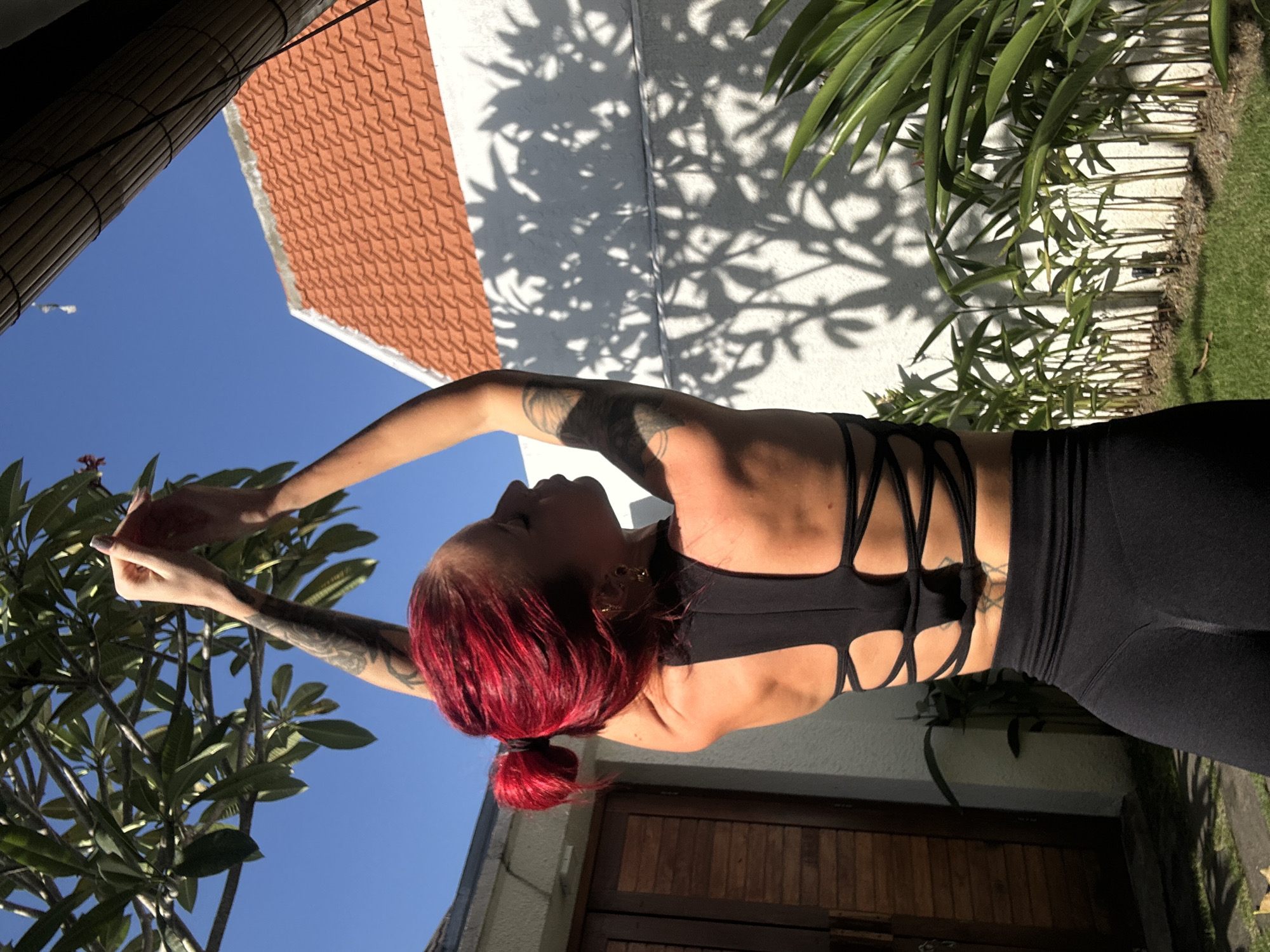 A woman with red hair pulled back in a ponytail stands outside in North Kuta, Indonesia, wearing black workout clothes. She is reaching up towards the leaves of a tree under a clear blue sky. Behind her is a white wall with shadows of leaves, a brown tiled roof, a closed wooden door, and a woven bamboo screen. Green foliage and grass are visible on the right.