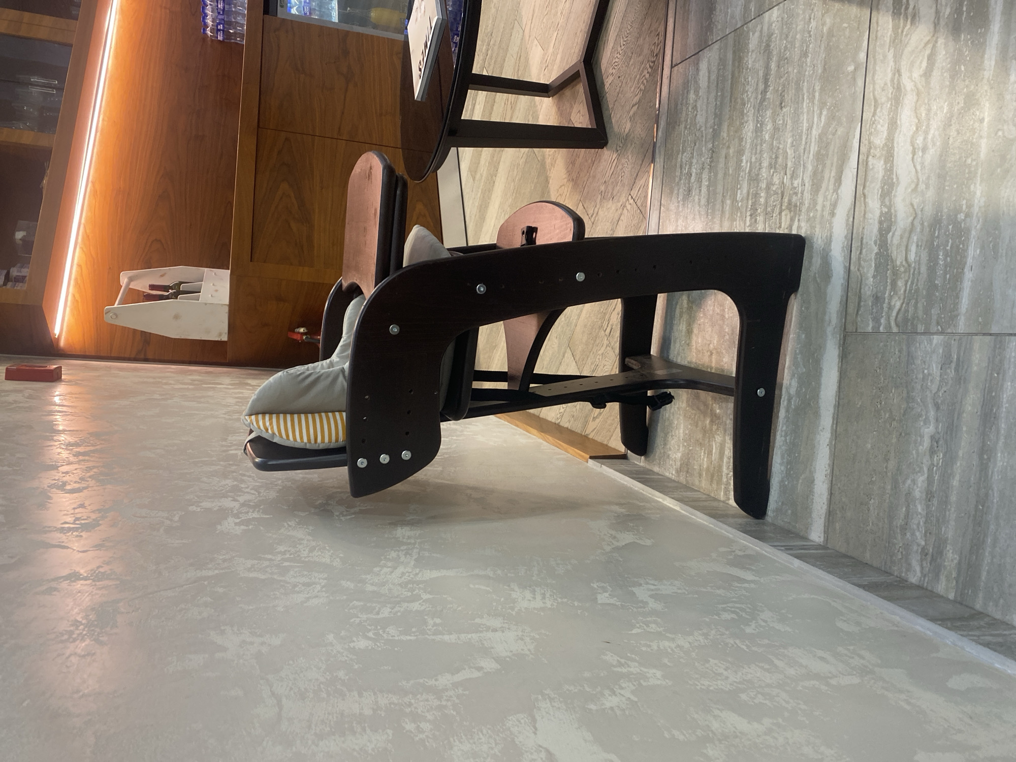 An indoor scene with a dark wood high chair situated next to a coffee table. The high chair has a light-colored cushion with orange and white stripes. The coffee table is round and dark, with a book on its surface. Behind the high chair, a wall with a textured, off-white finish is visible. Further in the background, there is a wooden cabinet with shelves and bottles of water. The floor is tiled with a pattern resembling marble. The overall setting appears to be a cafe or restaurant.