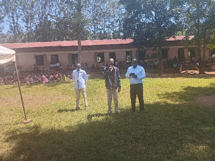 The image displays an outdoor daytime scene in Idakho South ward, Kenya, featuring a gathering on a grassy field in front of a building. In the mid-ground, three adult males stand erect. The central male, dressed in a dark jacket and tan trousers, holds a dark microphone-like object to his mouth, suggesting he is speaking. To his immediate left, a male in a light long-sleeved shirt and tan trousers stands with his hands clasped behind his back. To the right of the central figure, a third male in a light blue long-sleeved shirt and dark trousers observes a dark rectangular device held in both hands. On the far left of the frame, a light-colored pole with a circular base is partially visible, indicating a tent or flag structure.
In the background, a long, single-story building with a reddish-brown corrugated metal roof and light-brown, possibly unpainted, walls serves as the backdrop. The building features multiple window openings, some appearing unglazed, and several doorways. In front of and around this building, a large group of individuals, primarily children or students, are seated on the grass and on a slightly elevated platform or veranda. Many are wearing white shirts, potentially uniforms, interspersed with various other colored garments. Behind the building, a dense row of tall, slender trees lines the horizon, contrasting with a clear blue sky. The scene is brightly lit by direct sunlight, casting distinct shadows on the grass.