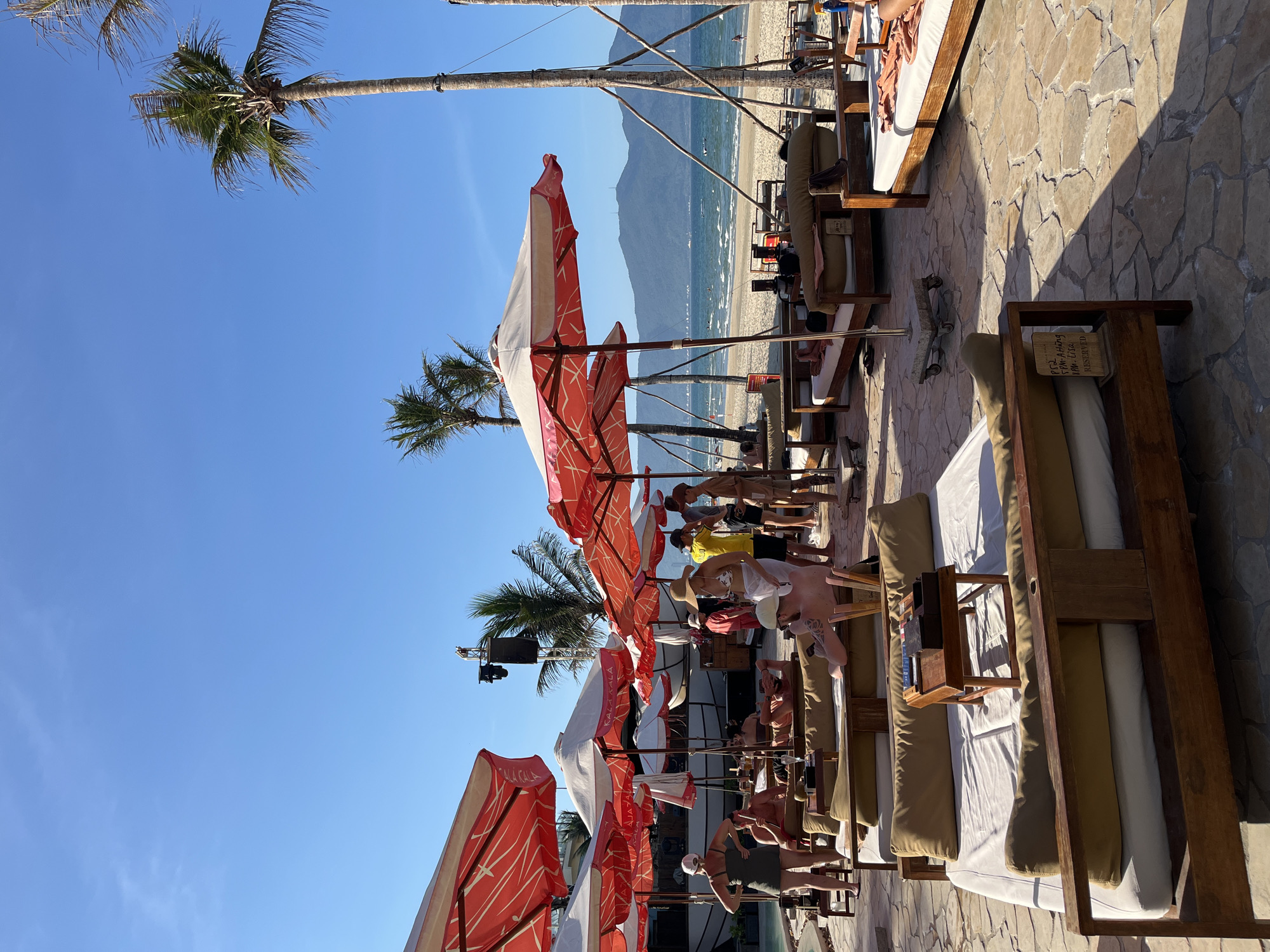 The image depicts a beach scene in Đà Nẵng, Vietnam. Palm trees and red-and-white striped umbrellas line the beach. Several people are on lounge chairs, some appear to be sunbathing. In the background, there is a mountain range across the water. The sky is clear and blue. The foreground shows a stone paved area with wooden framed lounge beds. Some beds have reserved signs. A large speaker is present. Some of the people are standing by the edge of the paved area and talking.