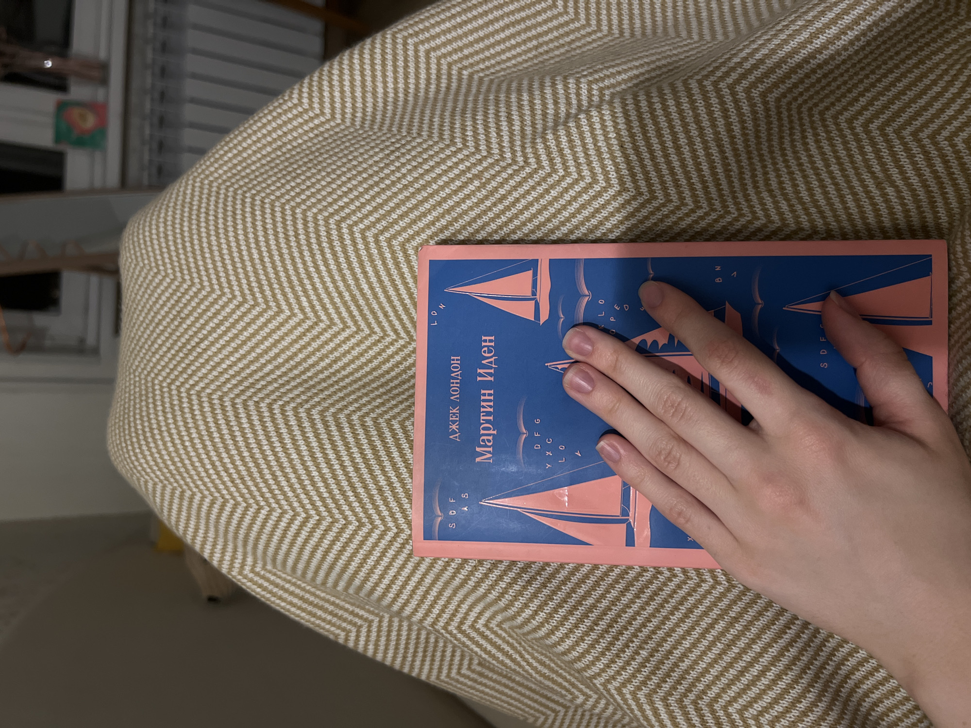 A hand rests on a copy of Jack London's "Martin Eden" in Russian, centered on a surface with a chevron pattern. The setting appears to be indoors, inferred from the chair or ottoman on which the book lies and the faint presence of a window or shelf unit in the background. The exact location within Saint Petersburg is not discernible from the visual elements of the image.