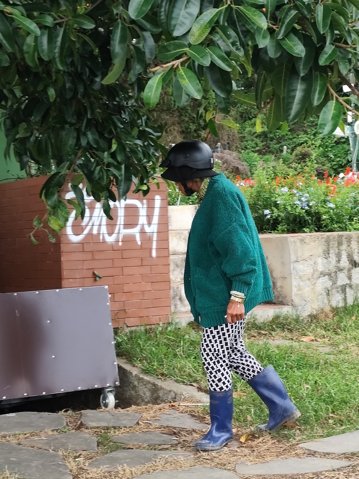 A person is walking outdoors in an area with cobblestone paths and grass, near a brick wall with graffiti and a metallic structure with wheels. They are wearing a black helmet, a green knitted cardigan, black and white patterned pants, and blue rubber boots. In the background, there is a wall with flowers and foliage. The setting appears to be a garden or park area within the city of Da Lat, Vietnam, characterized by its lush greenery and potentially cool climate, evidenced by the choice of clothing and footwear.