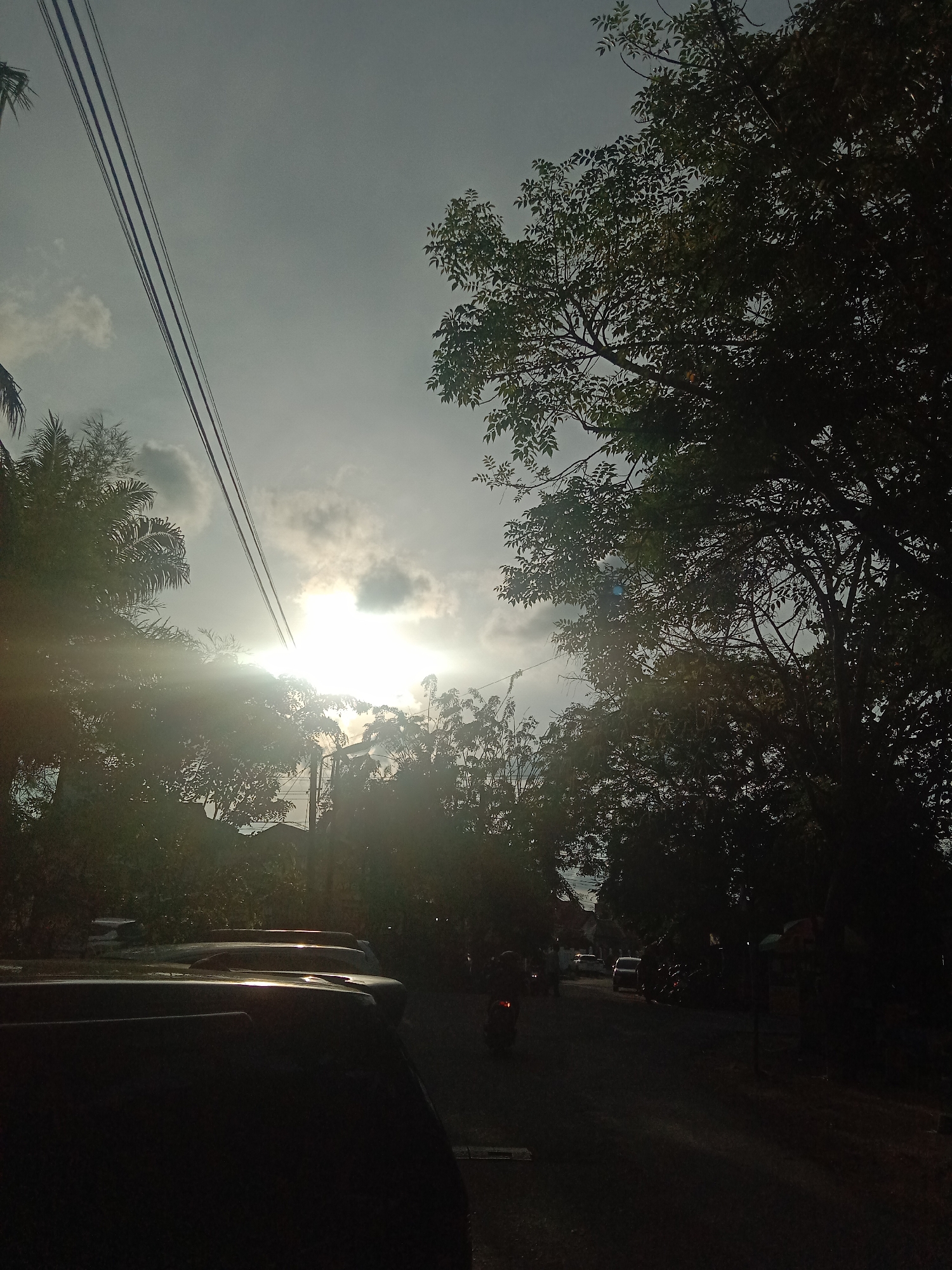 The image depicts a street scene in Lhokseumawe, Indonesia, captured during daylight hours. The sun is positioned behind clouds, creating a bright glare with sun rays piercing through the leaves of trees. The road appears dark, partially obscured by a row of parked cars in the foreground. In the middle distance, traffic includes a motorcycle and at least one car. Power lines run diagonally across the upper left of the frame. Trees flank both sides of the street, with varying degrees of visibility due to the strong backlighting. The overall scene has a high contrast, with dark shadows and bright highlights.