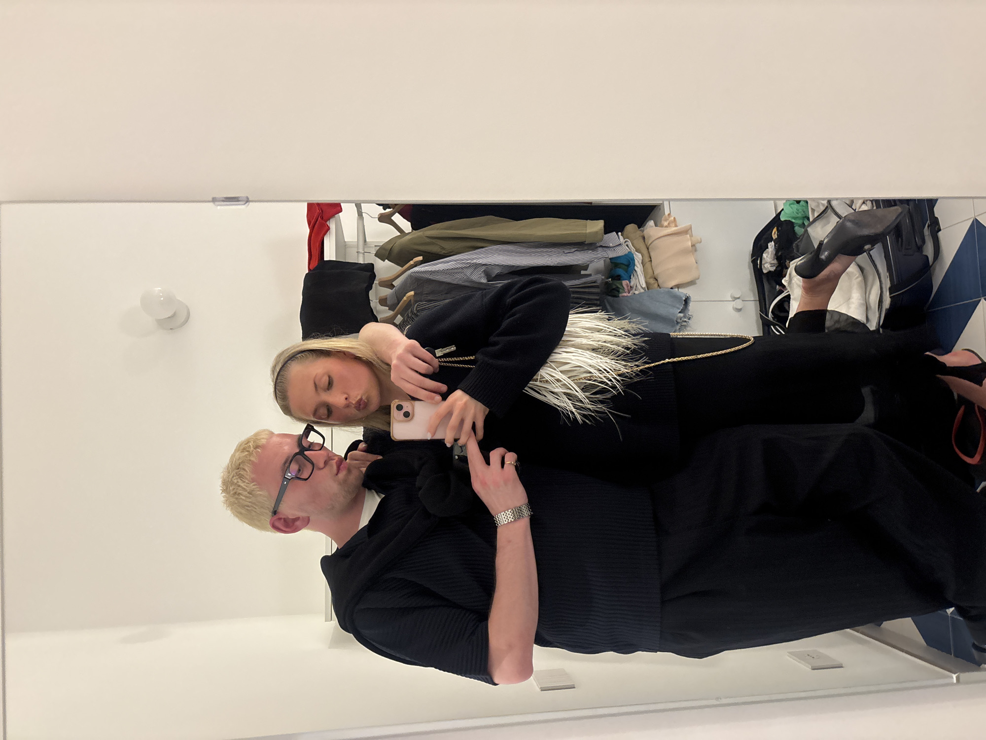 A photograph shows a reflection of a couple taking a selfie in a mirror, likely in a dressing room or closet. The man has short, dyed-blond hair, glasses, and is wearing a dark, textured shirt with short sleeves, and a silver watch on his left wrist. The woman is wearing a black top with white feather detailing, and black pants. She has a phone in her hands, seemingly taking the picture. The background shows a closet with clothes hanging on a rack, and some items on shelves. There is a light fixture on the wall. The flooring visible is blue and white tile. The location details are not discernible from the image, therefore Lipari, Italy cannot be confirmed.