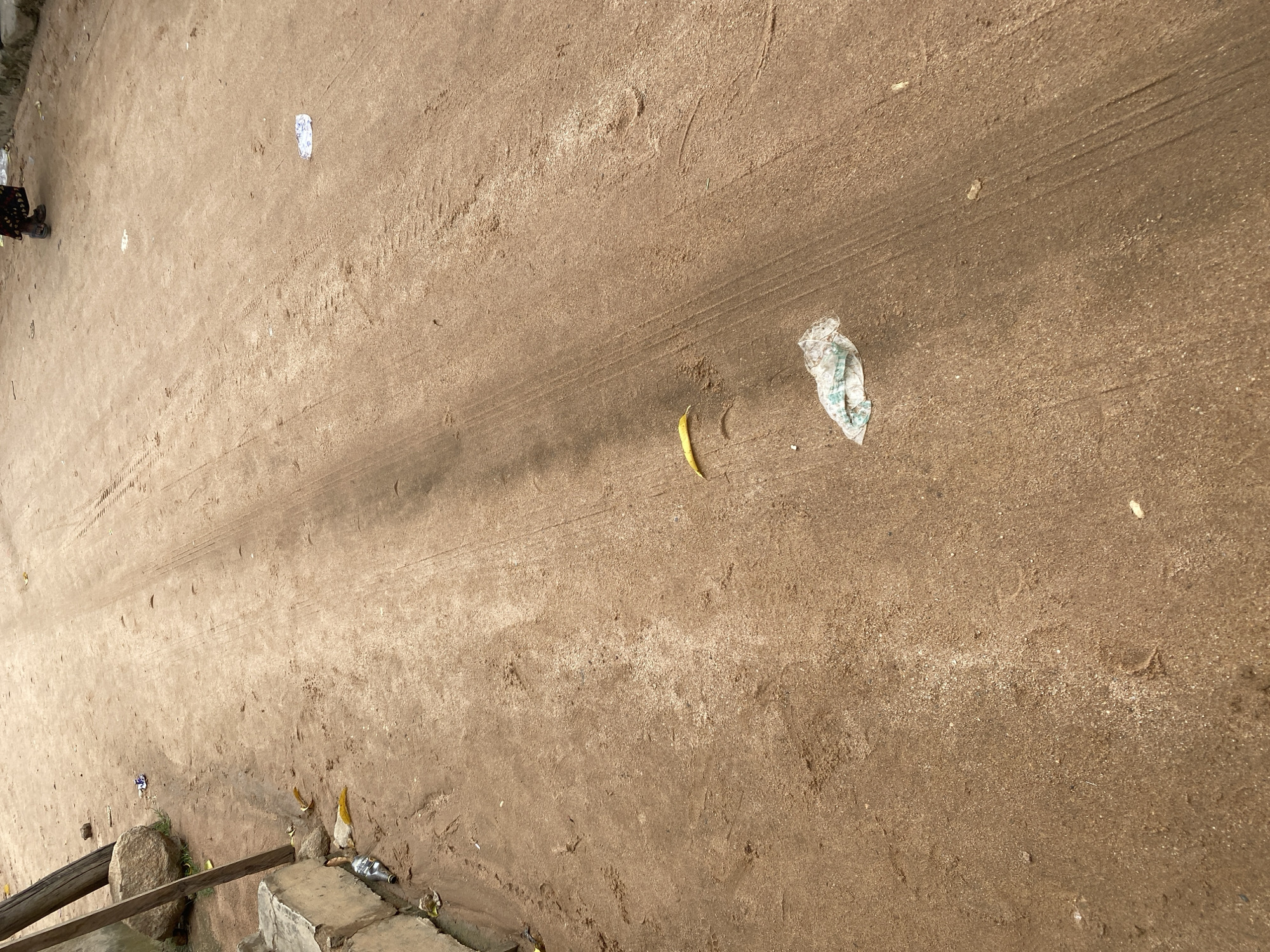 The image depicts a dirt road in Jalingo, Nigeria, characterized by a sandy texture and tire tracks. Litter is scattered across the surface, including plastic wrappers and a partially visible plastic bottle near a concrete barrier on the left side of the image. A few yellow leaves are also present. In the background, a person's legs and feet are visible, wearing dark sandals and a patterned skirt, walking away from the camera. Wooden poles and a large rock are situated near the concrete barrier. The overall scene suggests an unpaved urban or semi-urban environment with visible signs of human activity and waste.