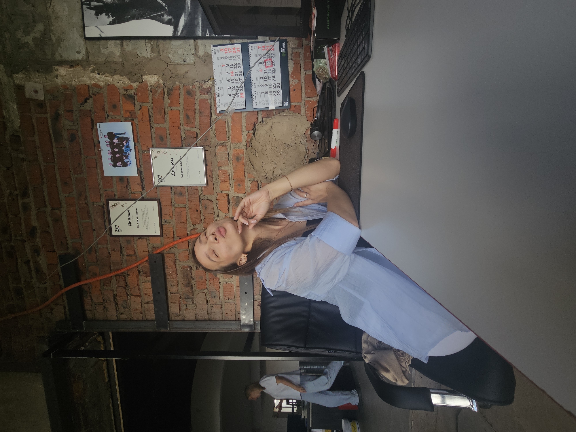 A young woman with long brown hair is sitting at a desk in an office, puckering her lips and resting her chin on her hand. She is wearing a light blue button-down shirt over a dark top. In the background, a person wearing a white t-shirt and light blue pants walks by. The office has exposed brick walls, and there are several framed certificates and a group photograph hanging on the wall. A calendar for May and June 2026 is visible on the wall. A computer monitor, keyboard, mouse, and a red marker are on the desk. The overall impression is of a casual office environment. This scene is likely in Chaikovsky, Russia, given the mention of the city.