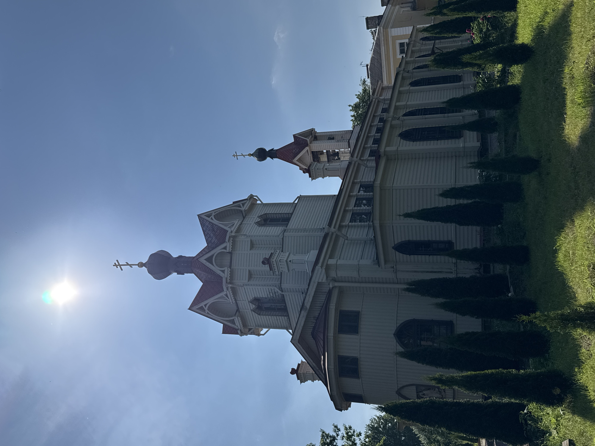 The image shows a white wooden church with red roofs and onion domes, viewed from a low angle against a bright blue sky. The sun is shining brightly, creating lens flare and casting shadows. The church has architectural details such as cross-topped domes, decorative eaves, and arched windows. In the foreground, a row of manicured evergreen trees lines a grassy area, suggesting a well-maintained churchyard. The scene appears serene and tranquil, likely captured on a clear, sunny day. The overall impression is one of religious architecture set within a peaceful, possibly historic, environment. Given the location context of Lomonosov, Russia, this is likely an Orthodox church. No people or specific events are depicted, and there is no visible text.