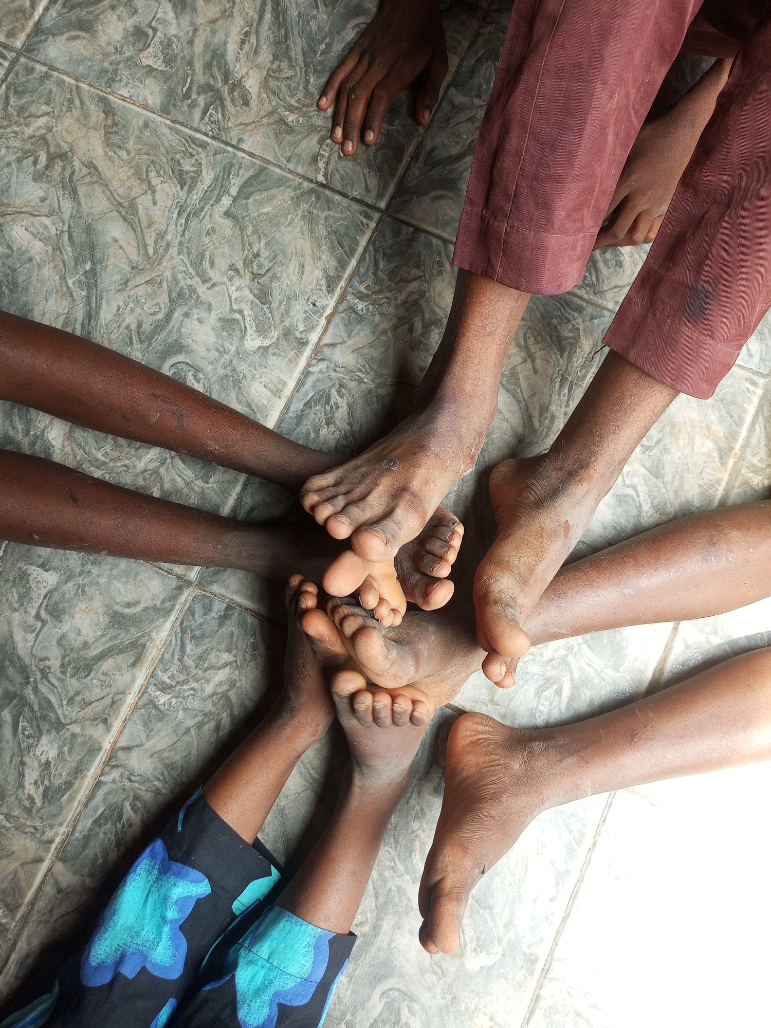 This is a top-down, close-up shot of several people's bare feet and legs on a tiled floor. The tiles are a light gray and white with a marbled pattern. In the foreground, a person wearing black pants with a blue and turquoise floral pattern has their legs and feet visible. To their right and slightly above, another person's legs, wearing reddish-brown pants, are visible, as well as their bare feet. More bare feet and lower legs are clustered in the center of the image, appearing to be touching or overlapping. One hand with dark skin and short fingernails is visible in the upper left corner of the frame, resting on the tiles. The lighting is bright, with a strong light source coming from the right, casting shadows. The scene depicts an informal gathering of people, possibly children given the size of some of the feet, in what appears to be a residential setting within Tempure, Nigeria.
