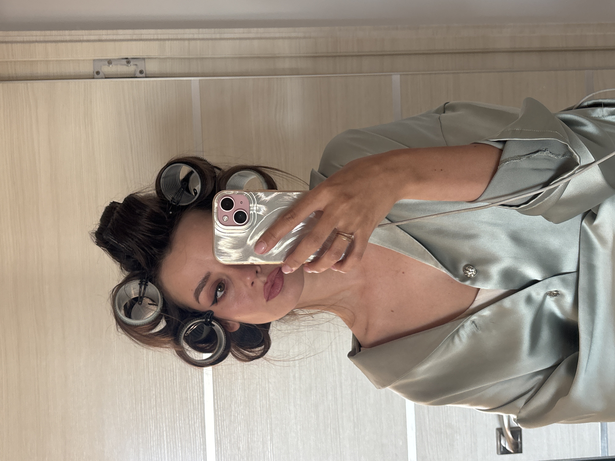 An adult female is depicted from the upper chest upward, taking a mirror selfie indoors in Krasnoyarsk, Russia. Her dark hair is styled with multiple dark hair rollers secured by clips. She wears winged eyeliner and lip makeup. A light green/sage-colored silky garment, possibly a robe or shirt, is worn over a lighter undergarment, featuring two decorative buttons. A metallic ring is visible on her right ring finger, and her right hand, displaying light pink nail polish on the thumbnail, holds a silver-colored smartphone with a dual-camera system (indicated by pink bezels), reflecting her image and the room. The background reflected in the mirror consists of light-colored, possibly wooden or laminate, wall and door surfaces, including a metallic door handle on the left and a metallic door hinge and frame on the right.