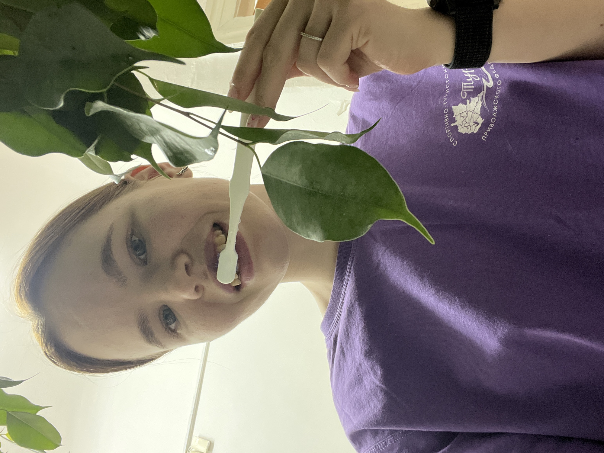 A low-angle, upward-facing shot shows a woman holding a ficus plant. She is fair-skinned with blue eyes and light brown hair pulled back. She wears a purple t-shirt with Russian text and an emblem depicting a river scene, possibly related to the city of Berezniki. A white, plastic-looking sap or secretion from the ficus plant is visible dangling from her mouth. She wears a black smart watch on her left wrist and a simple ring on her left ring finger. The background is white, suggesting an interior setting, potentially indoors in the city of Berezniki, Russia, given the shirt.