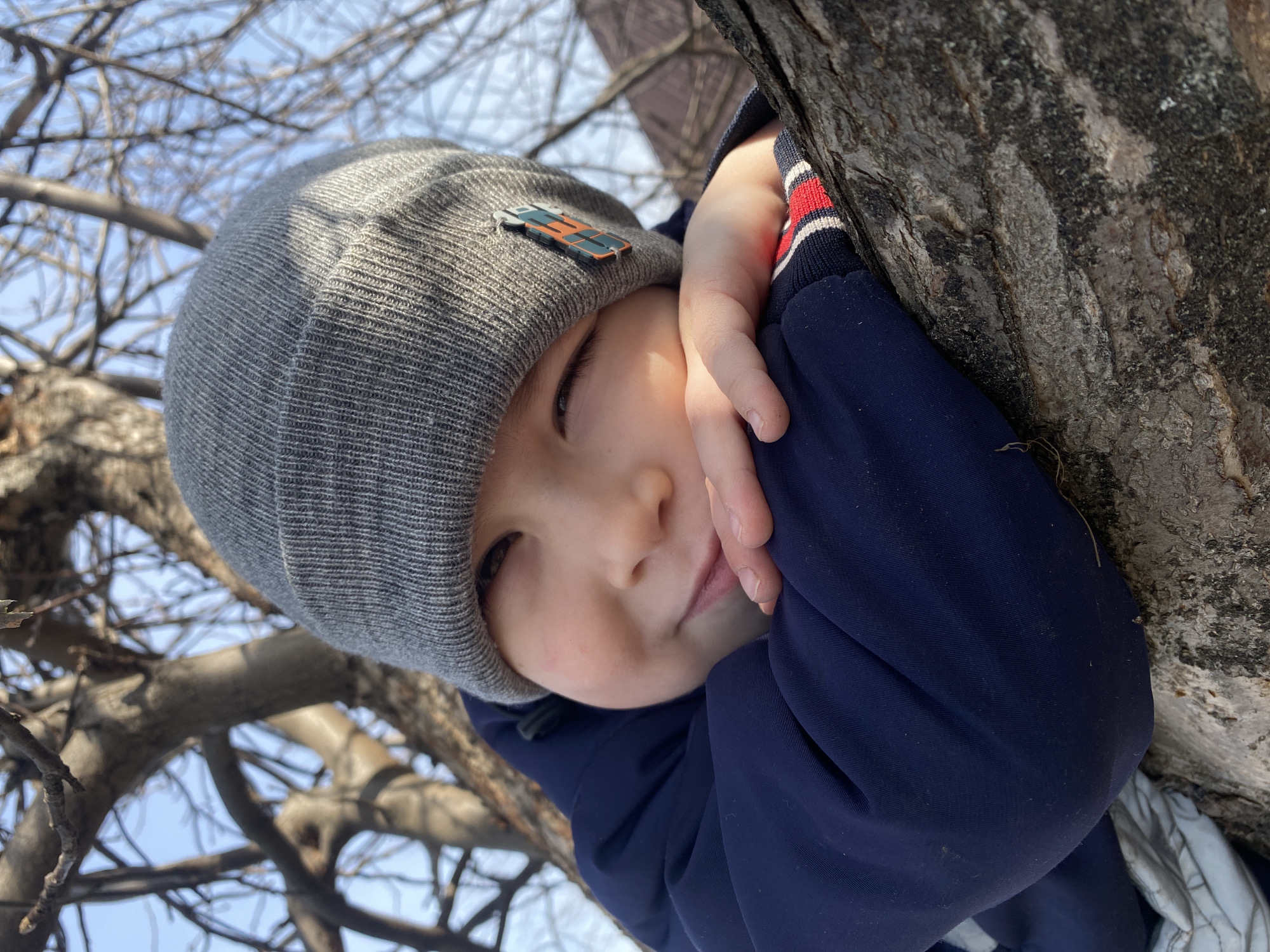 A young child wearing a gray beanie with an emblem on the front and a dark blue jacket is shown up close outdoors. The child is leaning against a tree trunk with their hand near their face. Bare branches of a tree are visible against a light blue sky in the background. The photo was taken in Ольховка, Russia.