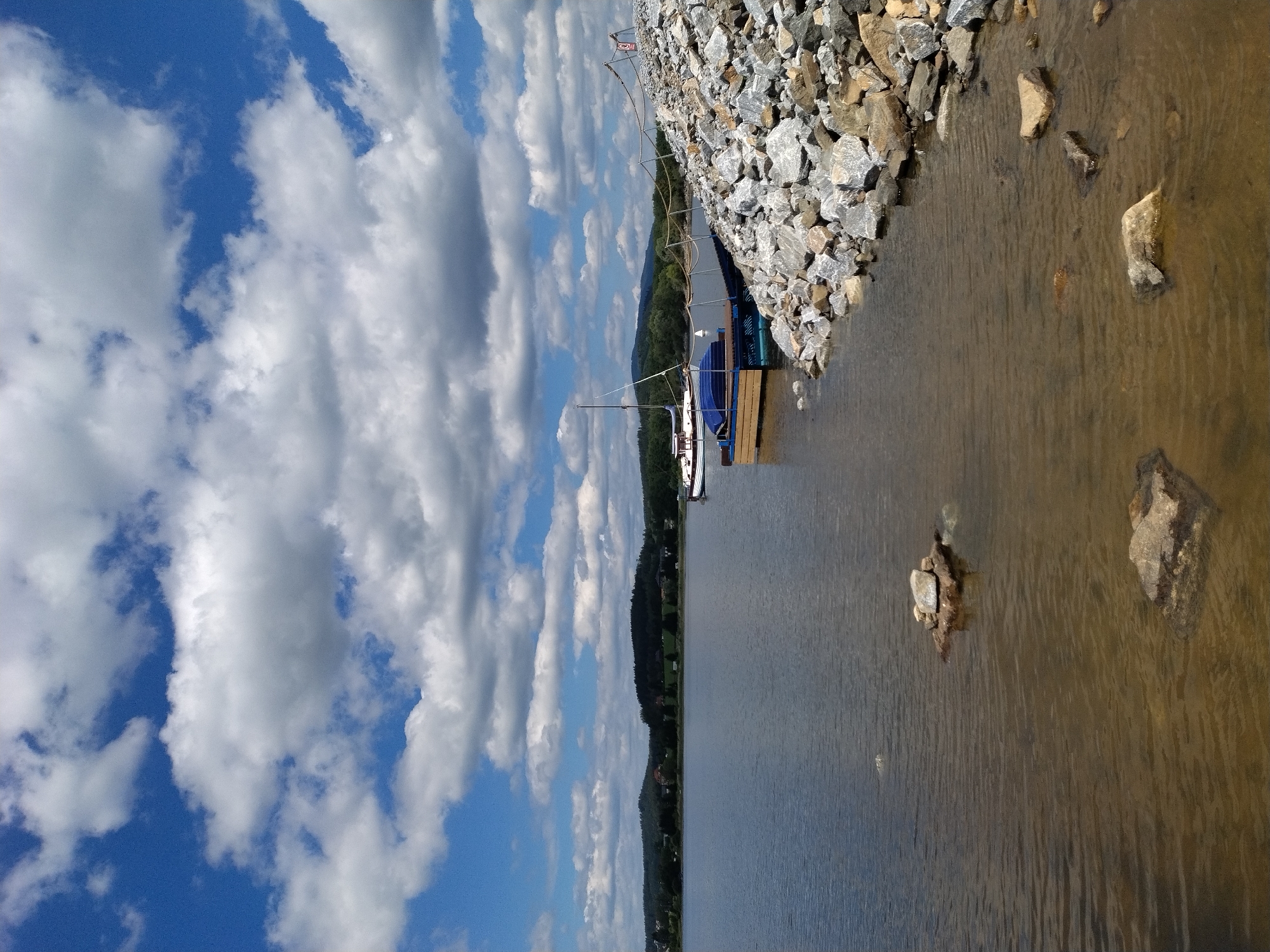 This is a vertical, medium-shot, exterior photograph taken on a sunny day with scattered clouds. The scene depicts a body of water, likely a lake, with a rocky shore on the right side of the frame. In the foreground, the shallow water shows several submerged rocks. A wooden platform with blue boat-like structures is moored to the shore, with a sailboat docked behind it. The background features a treed hillside and distant buildings. The sky is a vibrant blue with white, fluffy clouds. The image captures a tranquil, natural setting possibly near Radslav, Czechia.