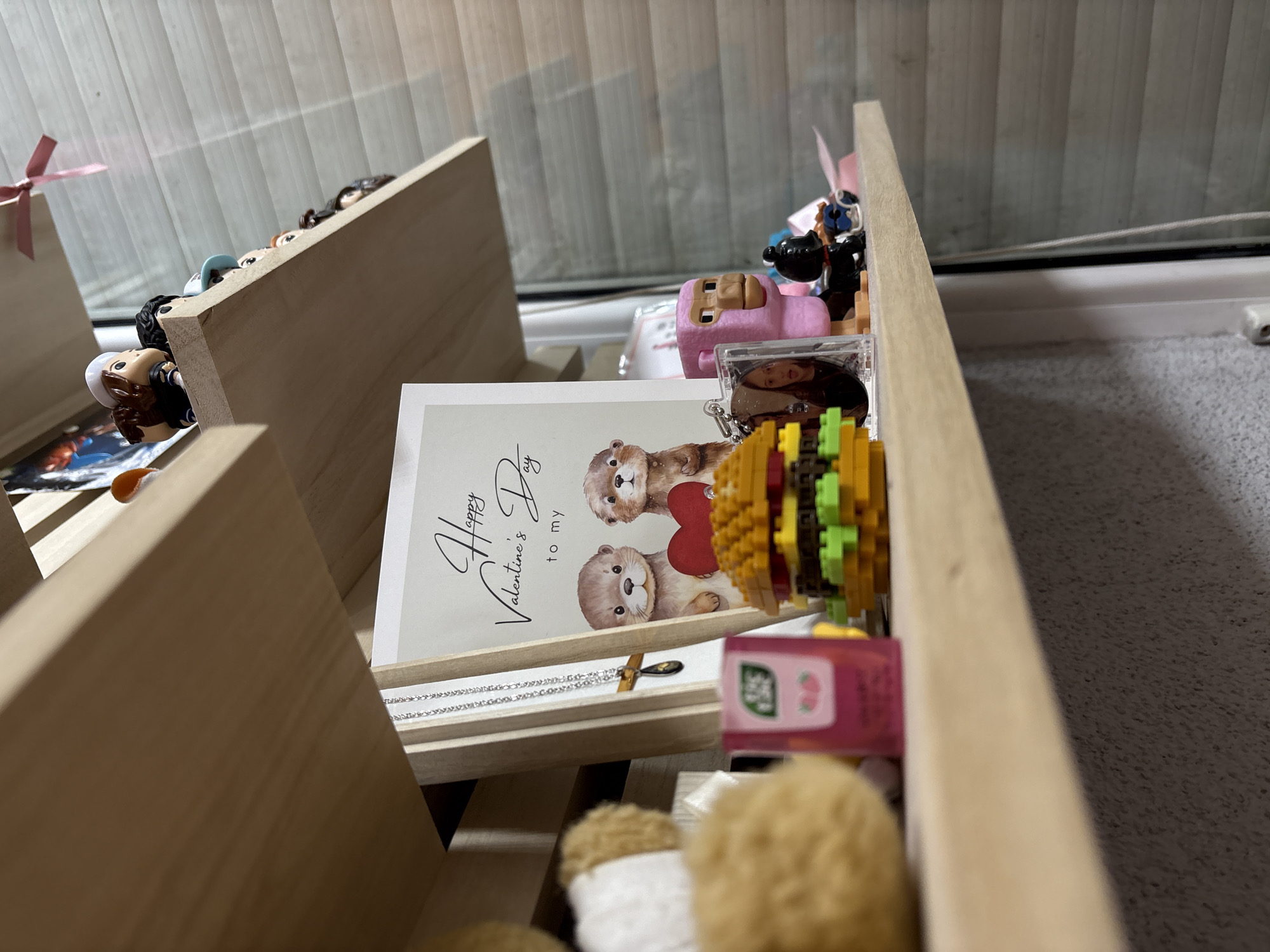 A close-up shot depicts a collection of objects placed on a tiered wooden shelf, likely located indoors near a window. The shelf contains various items: small figurines, including miniature people, a pink vehicle toy, and what appears to be a plastic burger. A Valentine's Day card displaying two otters holding a heart is prominently featured. A silver necklace is placed near the Valentine's Day card. Also there is a pink juice box. The exact location in Panagyurishte, Bulgaria, cannot be determined from the image.