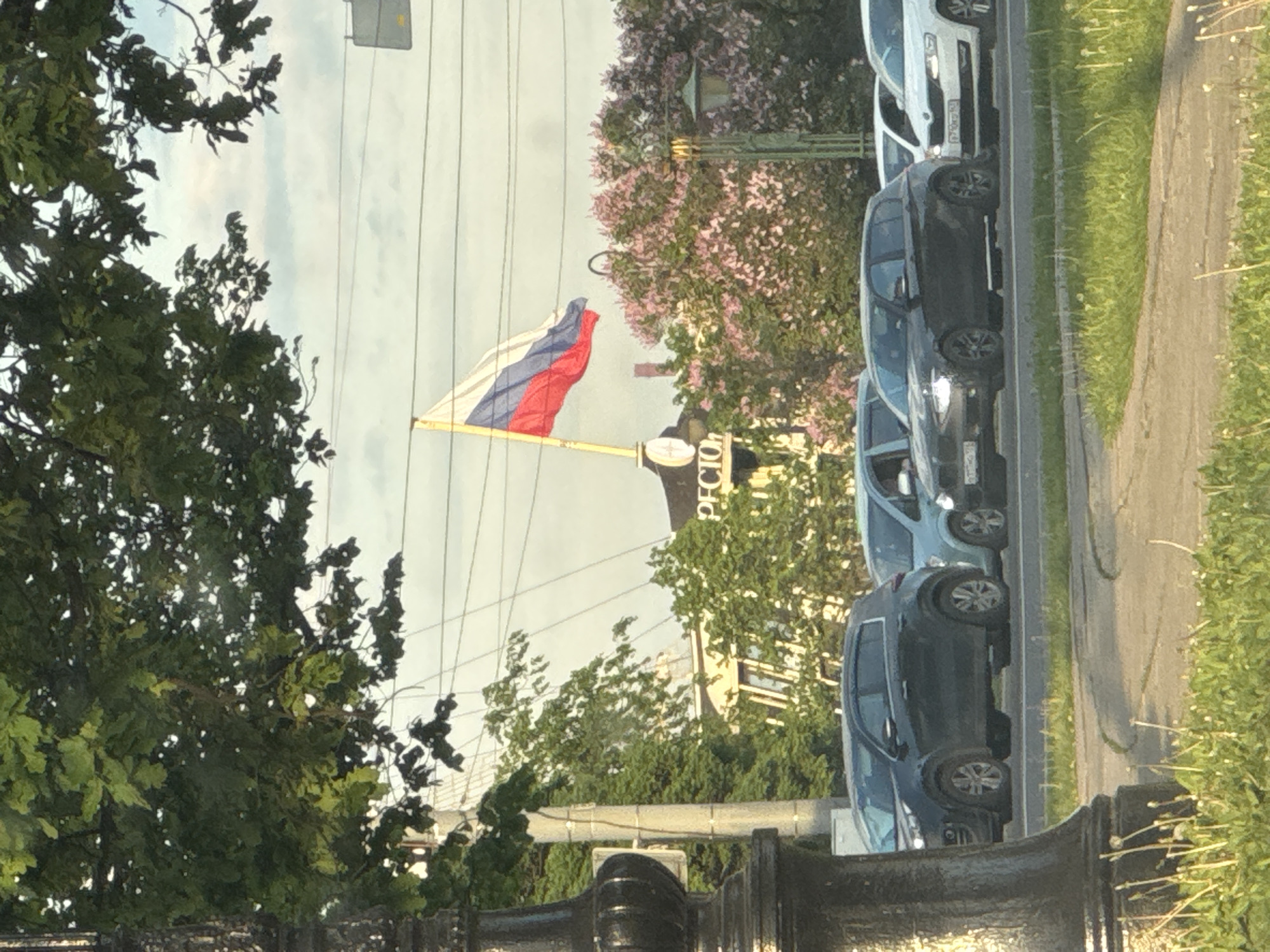 This daytime image, set in Saint Petersburg, Russia, captures an urban street scene under a bright, cloudy sky. A Russian flag, with its white, blue, and red horizontal stripes, is prominently displayed on a flagpole in the midground, gently waving. To the left, dense green foliage of trees, possibly oak, frames the view, while another tree adorned with pink or purple blossoms is visible behind the flag.
Along the right side of the frame, a row of cars is parked on the shoulder of a road, next to a grassy strip and a dirt path. Overhead utility wires crisscross the upper portion of the sky. In the background, partially obscured by trees, a building is visible, featuring a sign on which the Cyrillic letters "РЕСТОРА" can be discerned, indicating a "Restaurant" (Ресторан). The scene suggests a calm, typical day in an urban environment, likely in late spring or early summer given the tree blossoms.