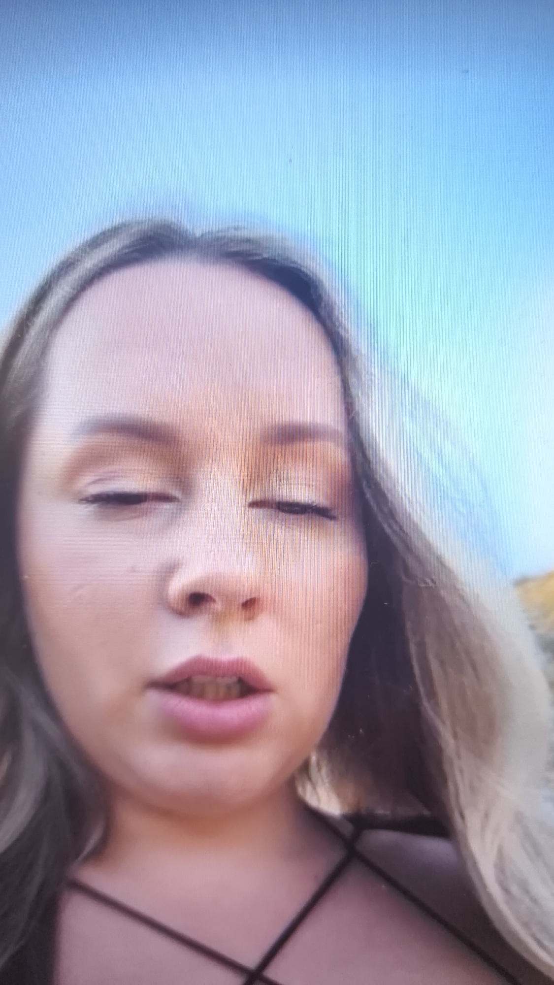 The media file presents a close-up portrait of a young woman, with her face and upper chest dominating the frame. She has light-colored, possibly blonde or light brown hair, visible on both sides of her face. Her eyes are partially closed, and her mouth is slightly open, revealing some teeth. She appears to be wearing eye makeup and a dark top with thin straps that cross on her chest.
The background consists of a bright, clear blue sky, suggesting an outdoor setting during the daytime. A faint lighter-colored area, possibly distant land or a structure, is visible on the far right horizon.
Due to her partially closed eyes, her emotional state is ambiguous; she could be blinking, feeling relaxed, or momentarily tired.
A notable characteristic of the image is the presence of distinct vertical lines across the entire frame, which are typical of a photograph or screenshot taken of a digital display, such as a television or monitor. No text is visible in the image.