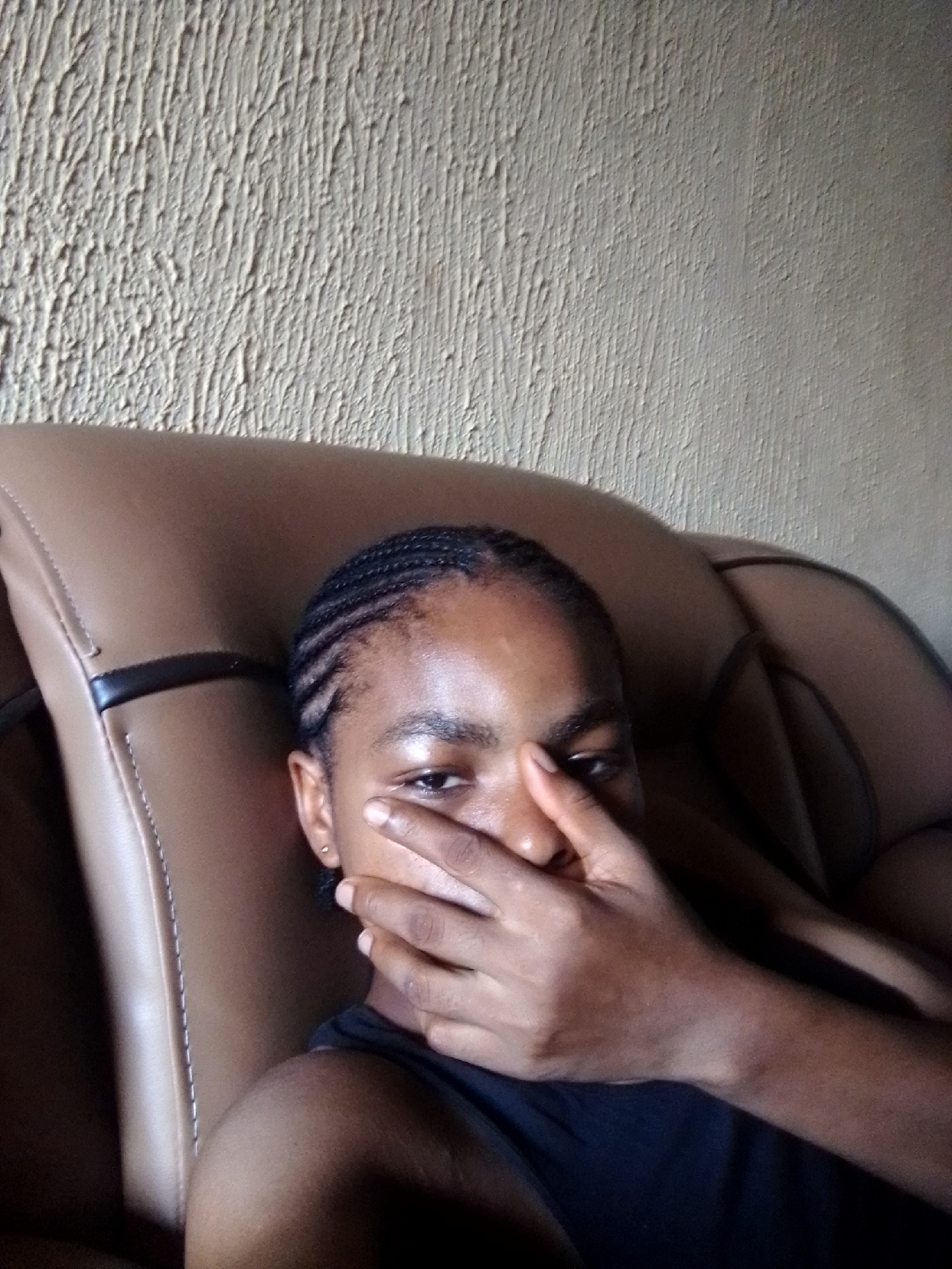 A medium shot depicts a young woman with dark skin and braided hair, seated in a brown leather chair in what appears to be Nkpor, Nigeria. She is wearing a dark-colored sleeveless top. Her right hand is covering the lower portion of her face, obscuring her nose and mouth. Her left eye is visible. The background features a textured, off-white wall. The chair is positioned near the wall, with a black accent stripe visible on the chair's armrest. The setting suggests an indoor environment, possibly a living room or similar space.