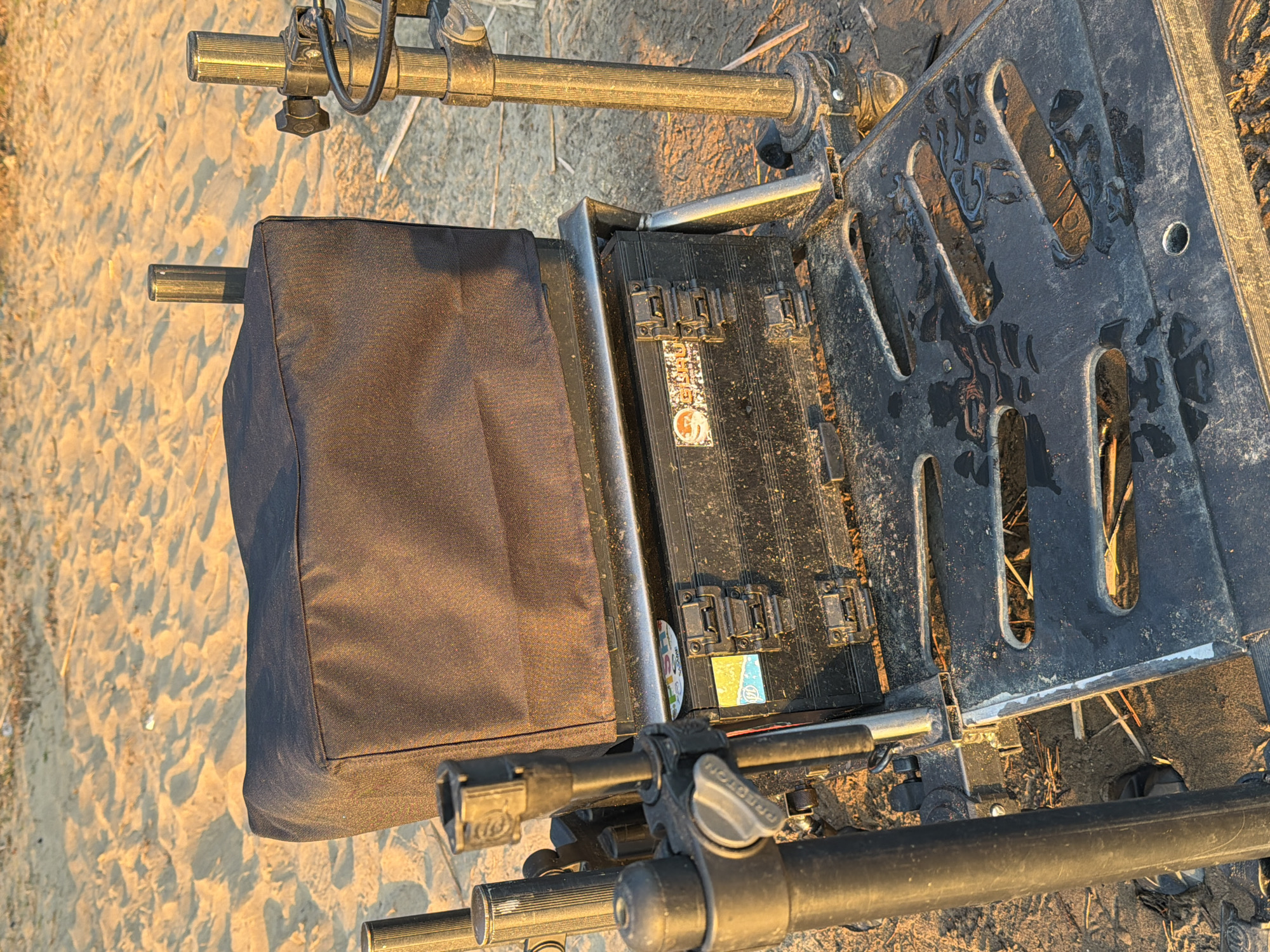 The image shows a piece of fishing equipment on sandy ground. The equipment includes a black seat box, a metal frame, and several adjustable rods. The seat box is covered in a dark blue or black fabric, and the frame appears to be made of a light-colored metal. There are several black plastic boxes attached to the frame. The location is likely near water, as indicated by the fishing equipment. The exact city of Chaykovsky, Russia is not discernible from the image.