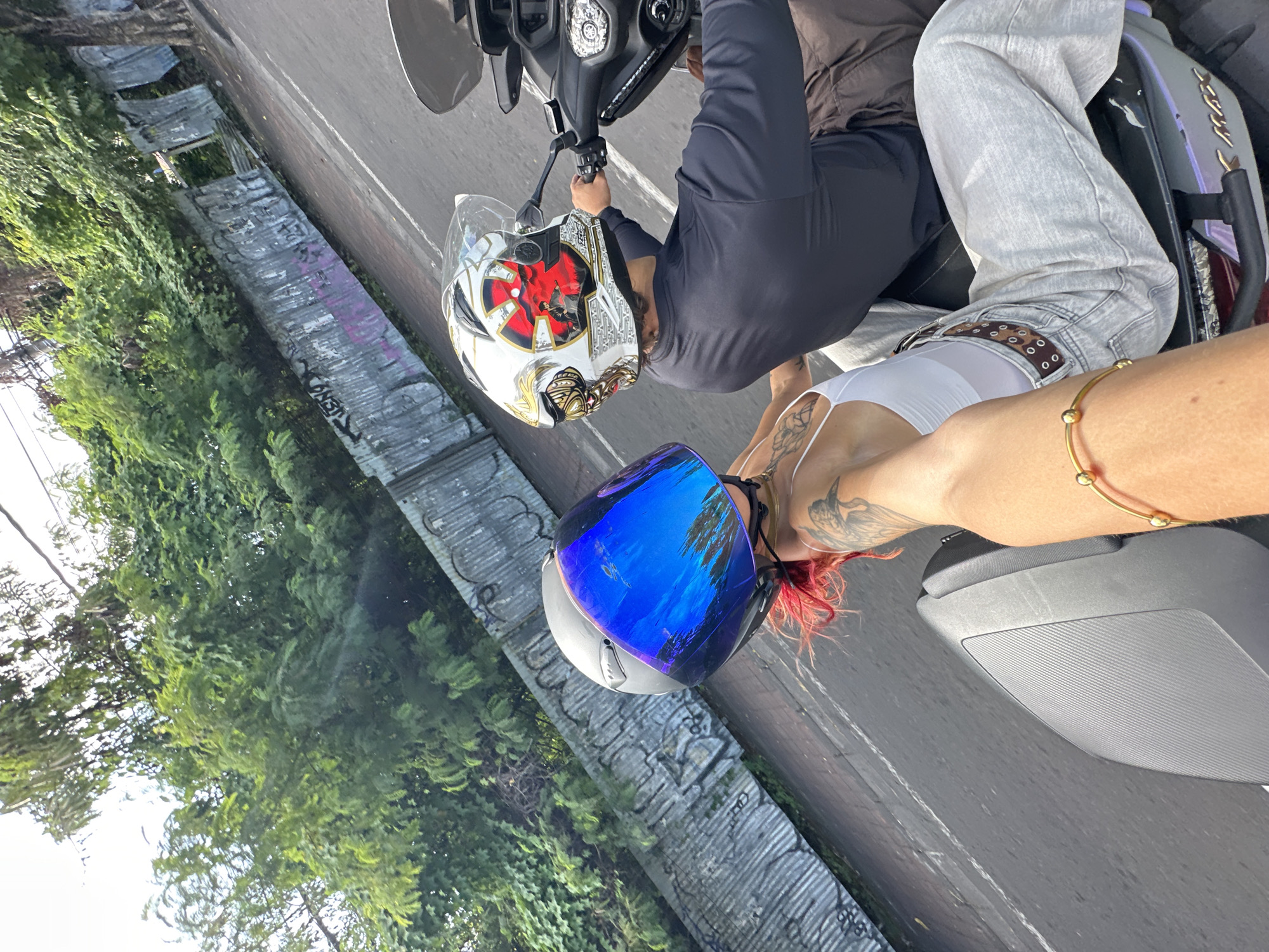 The image shows a scene with three individuals on a moving motorcycle in what appears to be a street in Kuta, Indonesia.
A person wearing a white and gold helmet with a red design is driving the motorcycle. Behind them sits another person wearing light-colored jeans, a black long-sleeved shirt, and a brown shirt, gripping the motorcycle.
A third person, presumably a passenger, is visible. They are wearing a white tank top and have red hair and several tattoos on their arm. They are wearing a blue and silver motorcycle helmet, too.
The street appears to be in a partly urban setting, with trees and a wall covered in graffiti alongside the road. The motorcycle is moving along the road.