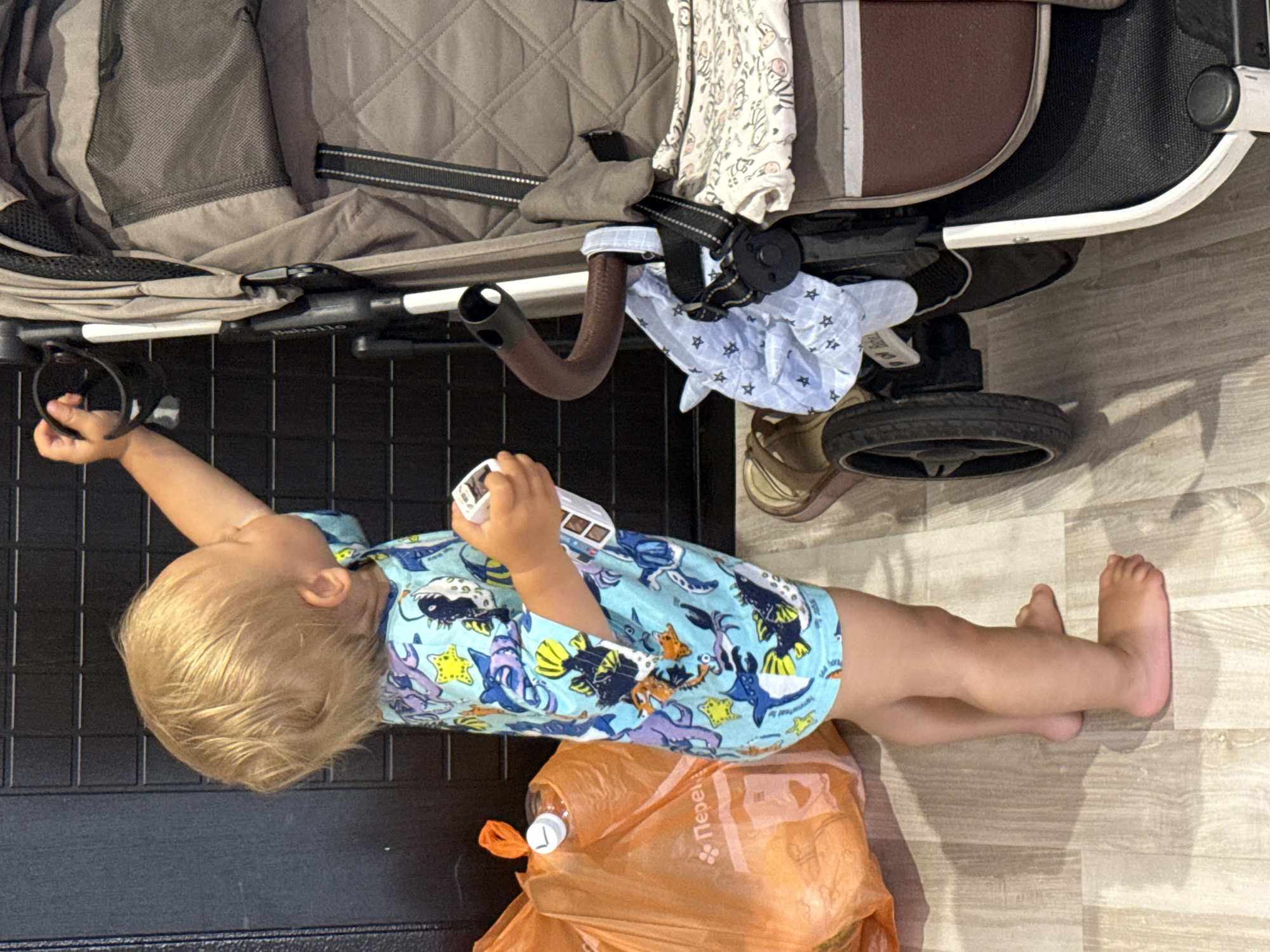 A young child with blonde hair and a light blue swimsuit with a sea creature pattern stands on a wooden floor. The child is reaching for the handle of a gray stroller, and is holding a small white toy car. Next to the child is an orange plastic bag filled with items, and a bottle with a white cap. The stroller has a brown handle and is partially covered with a light blue fabric decorated with stars and a white fabric with zebra prints. The background consists of a dark, textured wall. The scene appears to be indoors, possibly in a residential setting. There is no indication of the city of Sochi, Russia, in the image.