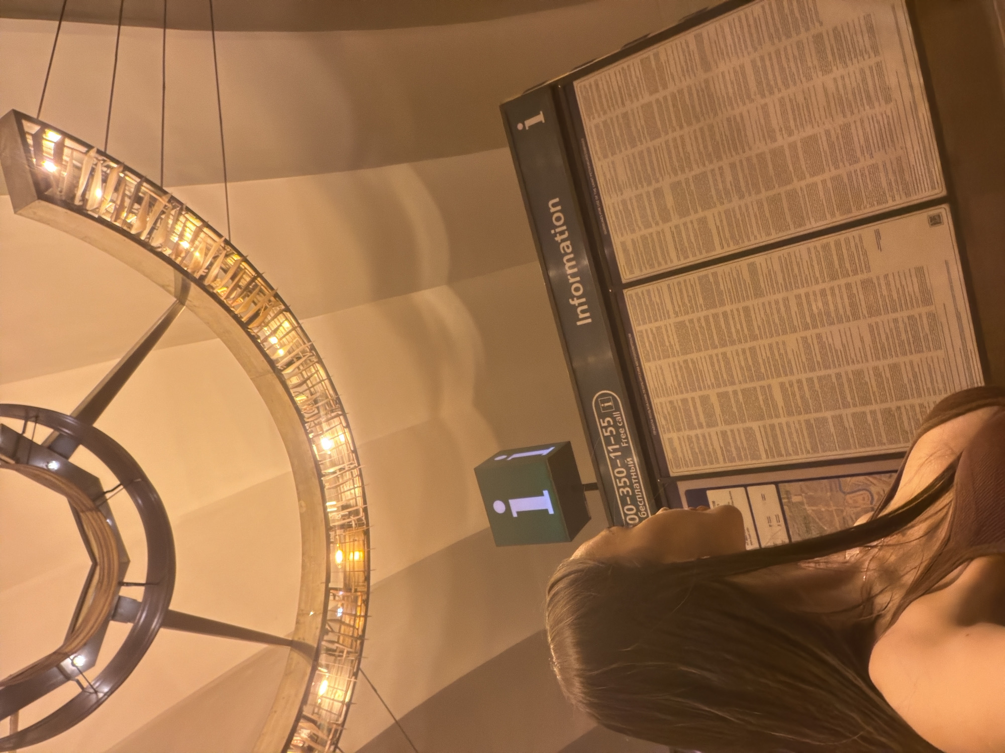 The image captures a woman from the chest up, looking to her right towards an information board in what appears to be a metro station in Saint Petersburg, Russia. The woman has long, dark hair and is wearing a dark top. She is standing in front of a large information display that contains a significant amount of text. Above the display, a sign reads "Information" and features an "i" icon. Another smaller, cube-shaped sign with an "i" icon is also visible. The background shows a domed ceiling with decorative lighting fixtures, typical of some Saint Petersburg metro stations. The overall lighting is warm and ambient.