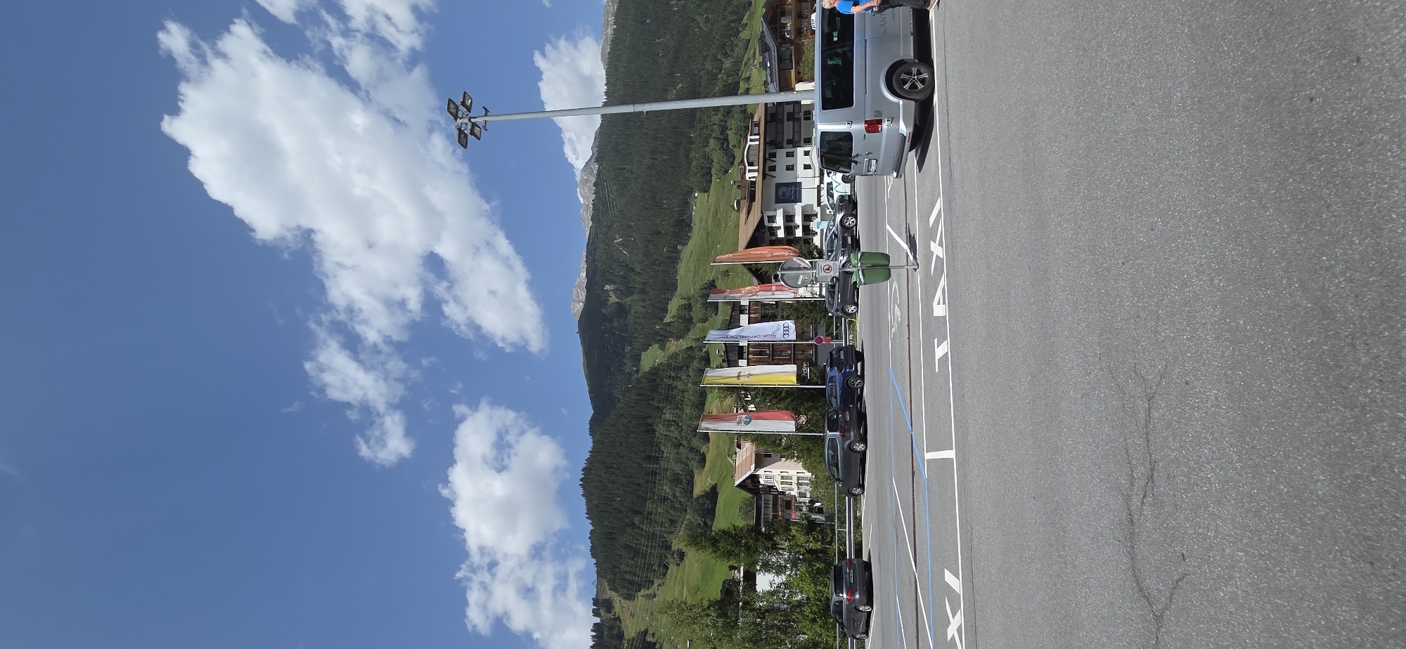 The image depicts a sunny day in Sankt Jakob am Arlberg, Austria, with a clear blue sky dotted with white, fluffy clouds. In the foreground, a paved parking lot is visible, marked with white lines designating parking spaces, some of which are labeled with the word "TAXI" in large white letters. Several cars are parked in the lot, including a silver minivan, a dark gray SUV, and a dark blue SUV. A person wearing a blue shirt and light-colored shorts stands on the right side of the frame, near the silver minivan.
In the middle ground, a row of colorful flags on poles stands in front of a line of buildings that appear to be hotels or shops, characterized by their alpine architectural style with wooden balconies and pitched roofs. Behind the buildings, a lush green mountain slope rises, covered with dense coniferous trees. A ski slope is visible on the mountain, suggesting the location is a popular ski resort. In the distance, the rugged peaks of the Alps are visible, some capped with snow. A tall lamppost with multiple light fixtures stands to the right of the flags, illuminating the parking area. The overall scene conveys a sense of a serene mountain village on a bright summer day.