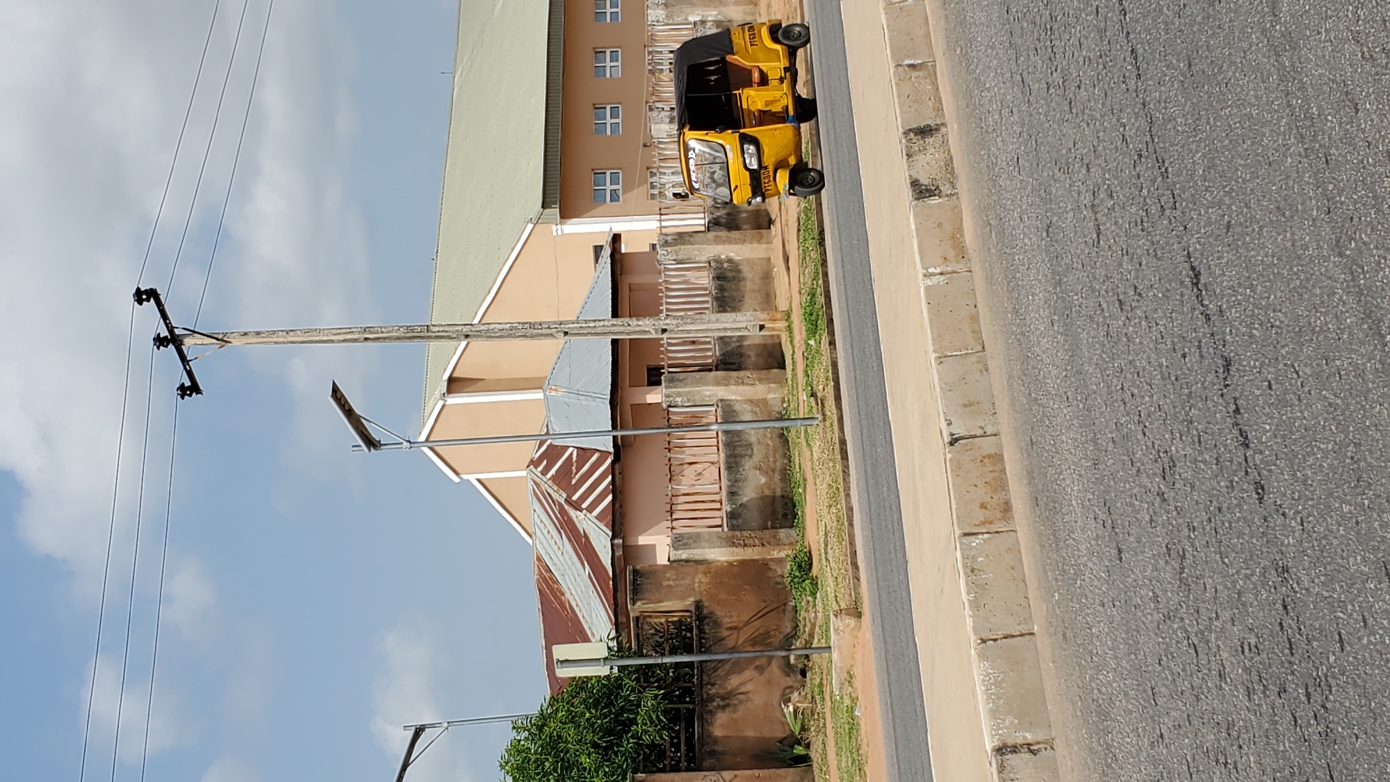 The image shows a street scene in Jalingo, Nigeria, with a yellow auto-rickshaw driving on the right side of the road. The auto-rickshaw has a black canopy and appears to be a mode of public transportation. In the background, there are buildings, including a house with a corrugated iron roof and a larger building with a lighter-colored roof and several windows. A utility pole with power lines and a solar-powered street lamp are also visible. The road is paved and bordered by a concrete curb and a strip of grass. The sky is partly cloudy, suggesting it is daytime. The overall scene depicts an ordinary urban or semi-urban environment in Nigeria.