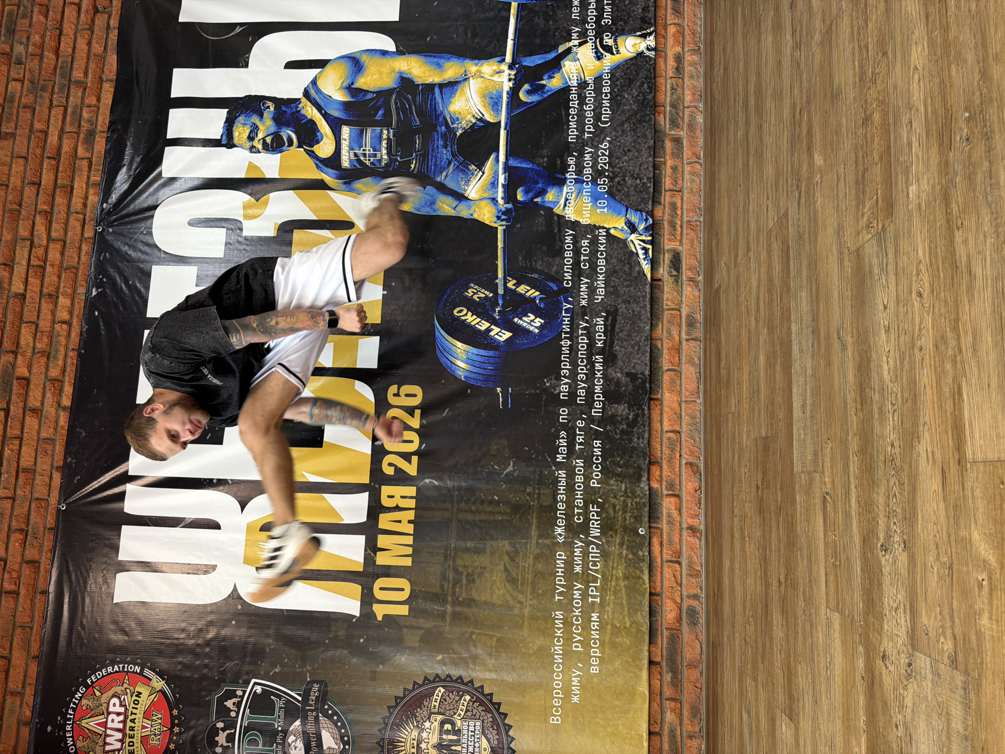 A man in athletic clothing stands in front of a banner advertising the "Iron May" All-Russian powerlifting tournament. The event, taking place on May 10, 2026, in Chaikovsky, Permsky region, Russia, includes powerlifting, squat, bench press, deadlift, power-sports, standing press, and bicep exercises. The banner features a picture of a weightlifter and logos for powerlifting federations. The banner is situated in front of a brick wall and a wooden floor.