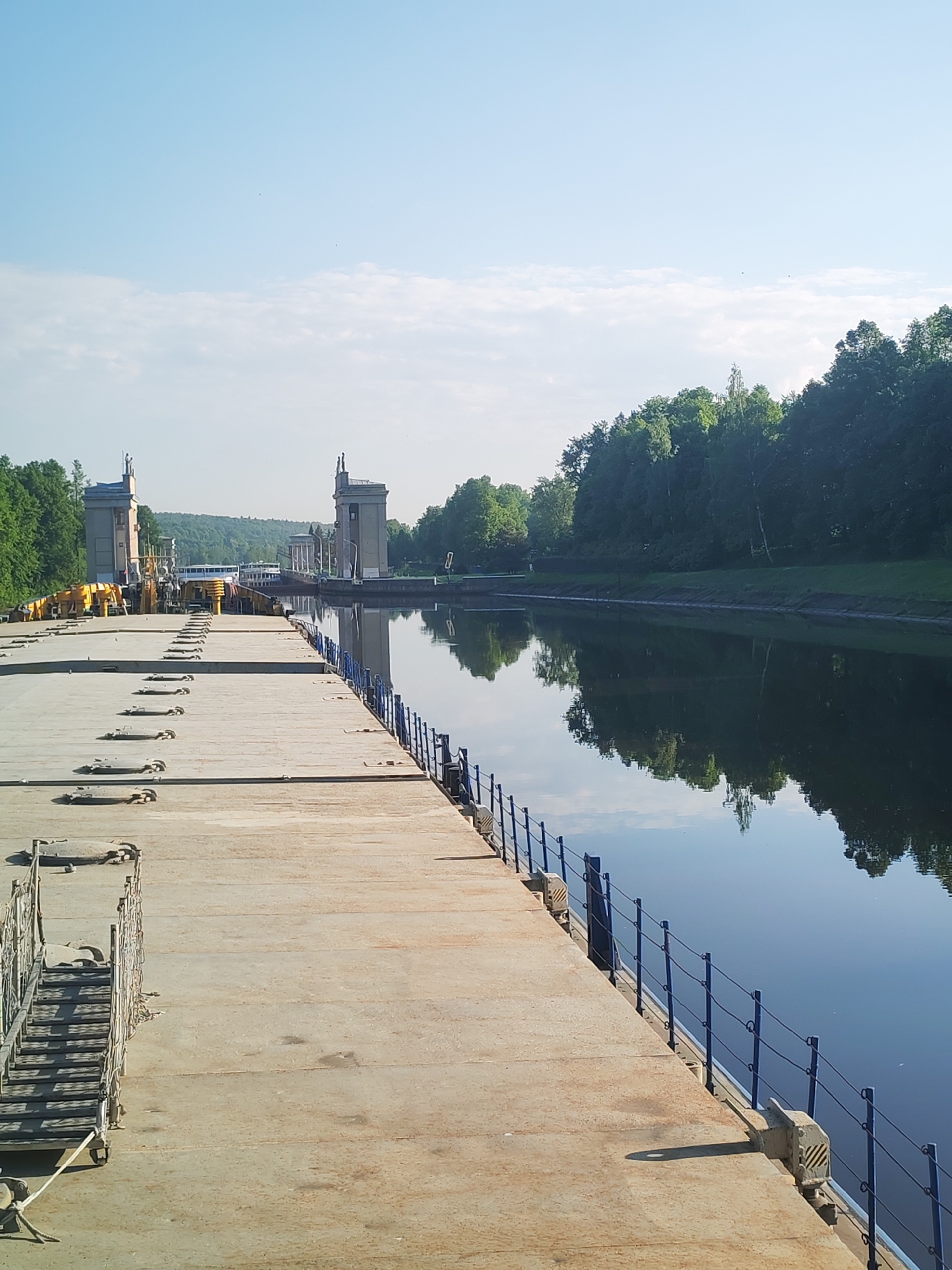 A photograph depicts a canal scene at the lock number 6 in Iksha, Russia. The foreground shows a concrete platform with metal tie-down points spaced along its surface. A portable staircase with wheels is at the front left of the platform. Along the water's edge is a blue-painted metal railing. The water reflects the sky and the tree-lined bank on the right. In the distance are two lock structures with sculptures atop. A vessel is visible within the lock. The sky is light blue with some high clouds.