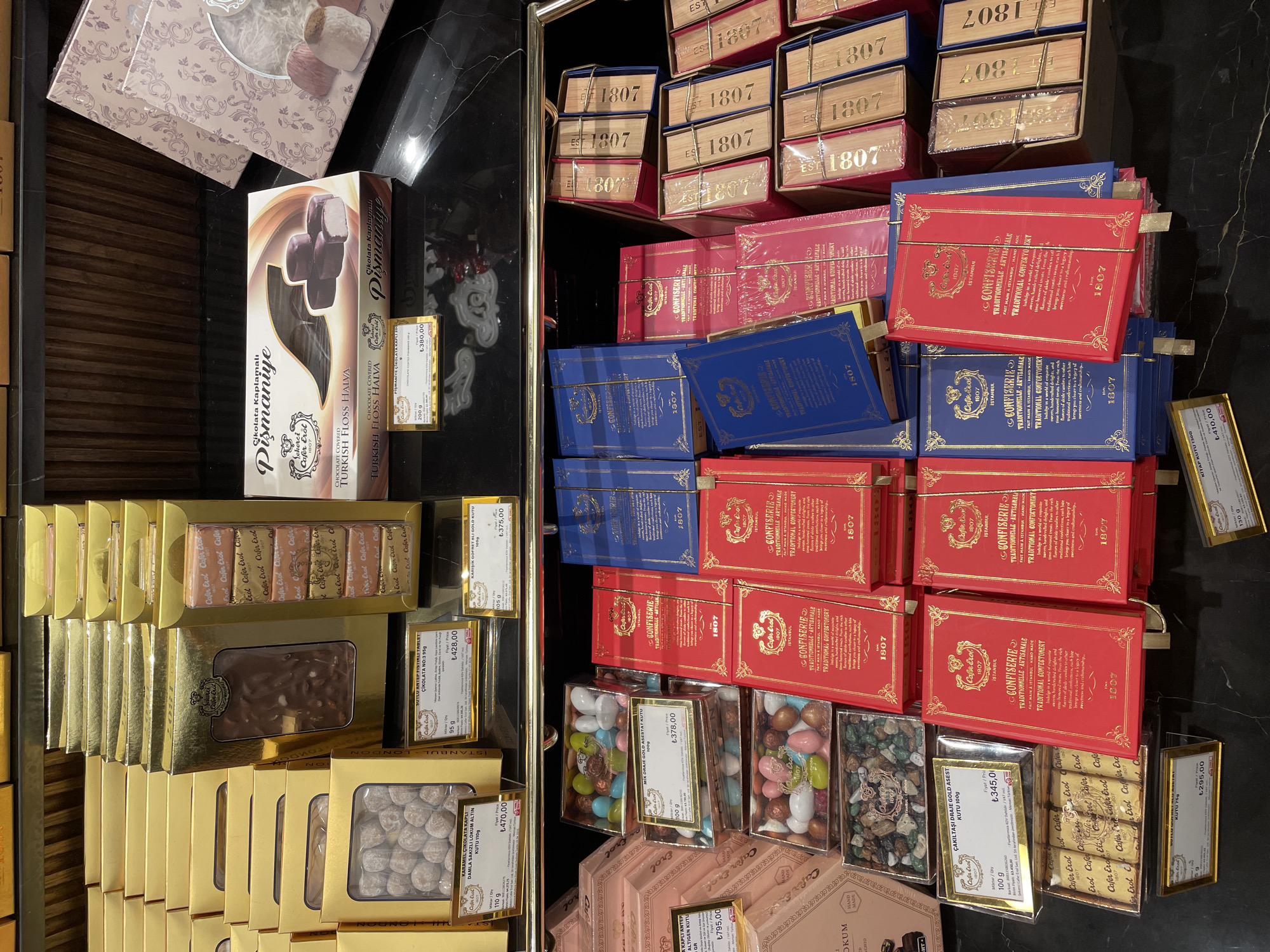 The image shows a display of assorted chocolates and confectioneries in a store. The products are arranged on shelves and in boxes, with gold, pink, blue, and red packaging. Many of the boxes have the brand name "Café Érol" and "Istanbul" printed on them, along with the year "1807". Some of the products are labeled in Turkish and English, with prices indicated in Turkish Lira. The items include chocolate bars, Turkish floss halva coated in chocolate, and mixed nut and dried fruit confections. The store appears to be well-lit with a clean, organized presentation of goods, typical of a high-end confectionery shop. The setting is likely a retail environment, possibly a specialty food store or a luxury gift shop, given the presentation and branding. Based on the visible branding and common luxury confectionery brands, it is plausible that this store is located in Kadıköy, a district on the Asian side of Istanbul, Turkey.